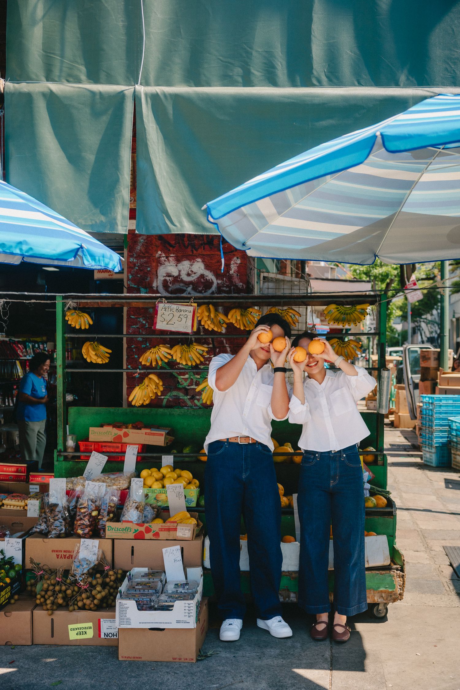 Couple holding oranges over their eyes at a vibrant outdoor fruit market stand with blue striped umbrellas.