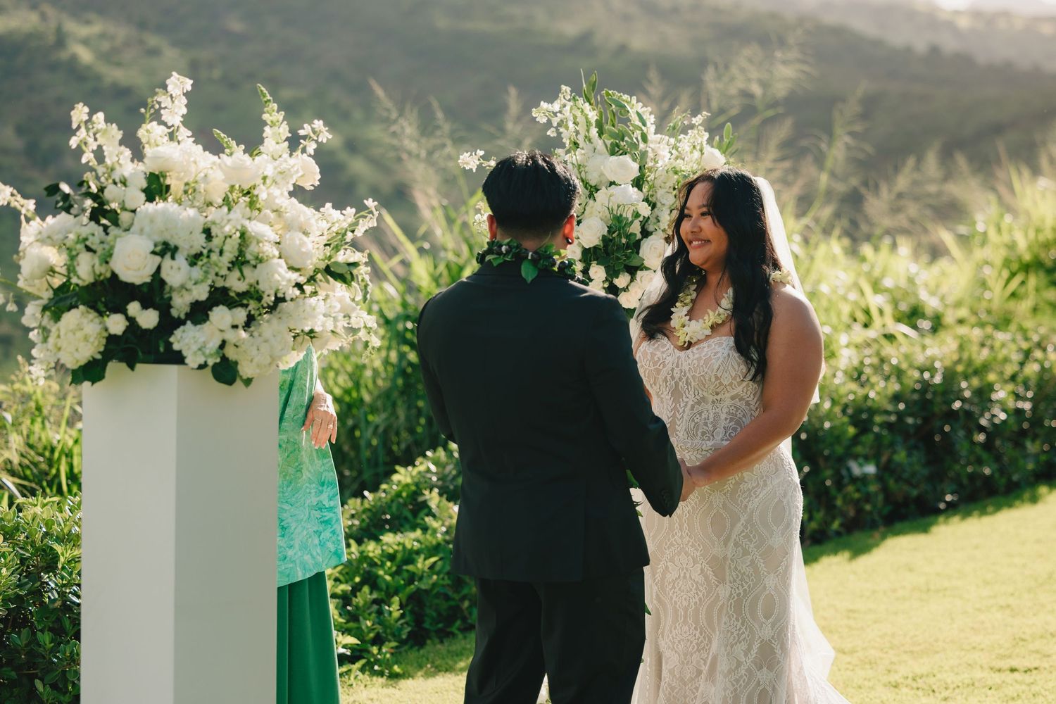 Couple exchanging vows outdoors at elegant wedding ceremony with white floral arrangements and lush green mountain backdrop.