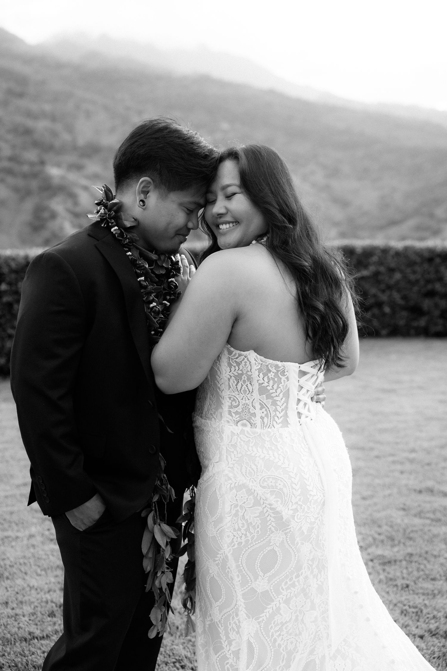 Black and white photo of a smiling bride in lace gown embracing groom wearing a floral lei at an outdoor wedding.