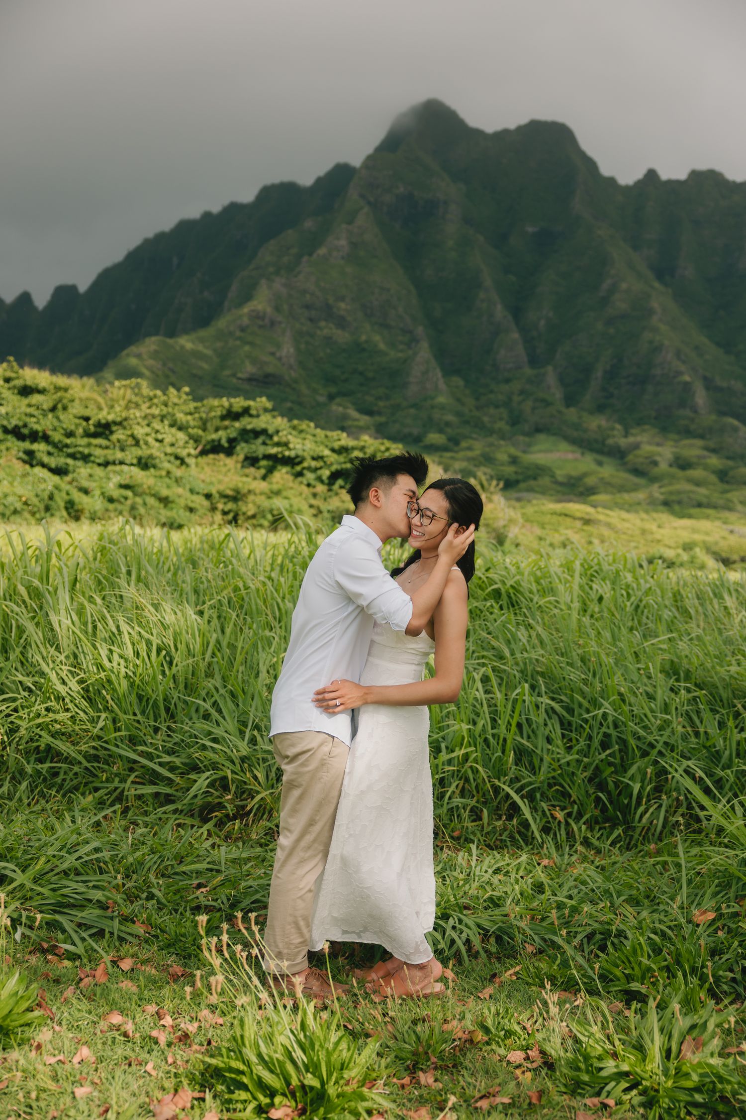 Couple sharing a romantic kiss in a lush green field with dramatic mountain backdrop in Hawaii.