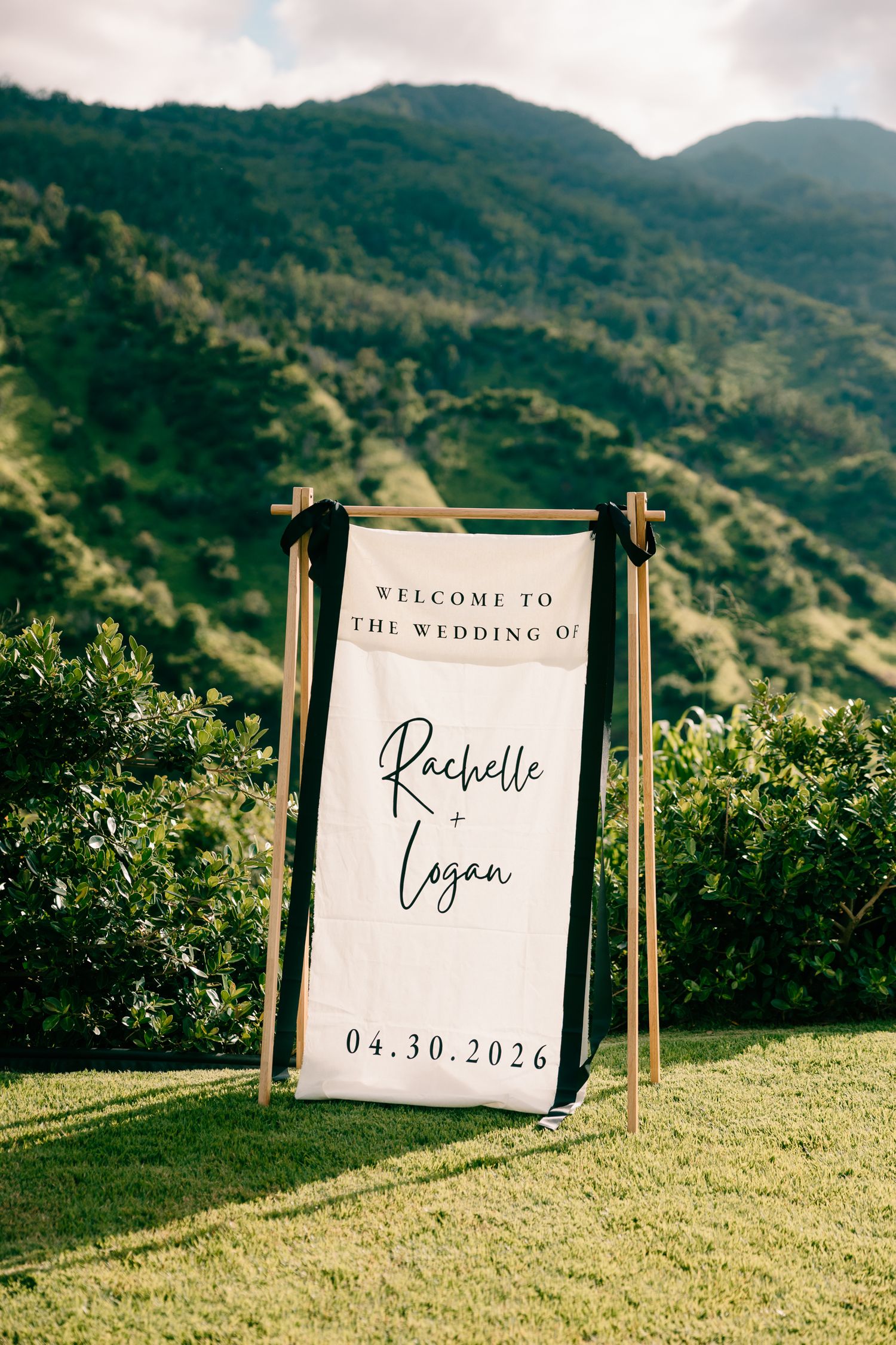 Outdoor wedding welcome sign for Rachelle and Logan on 04.30.2026, displayed on a wooden stand with lush green mountains.