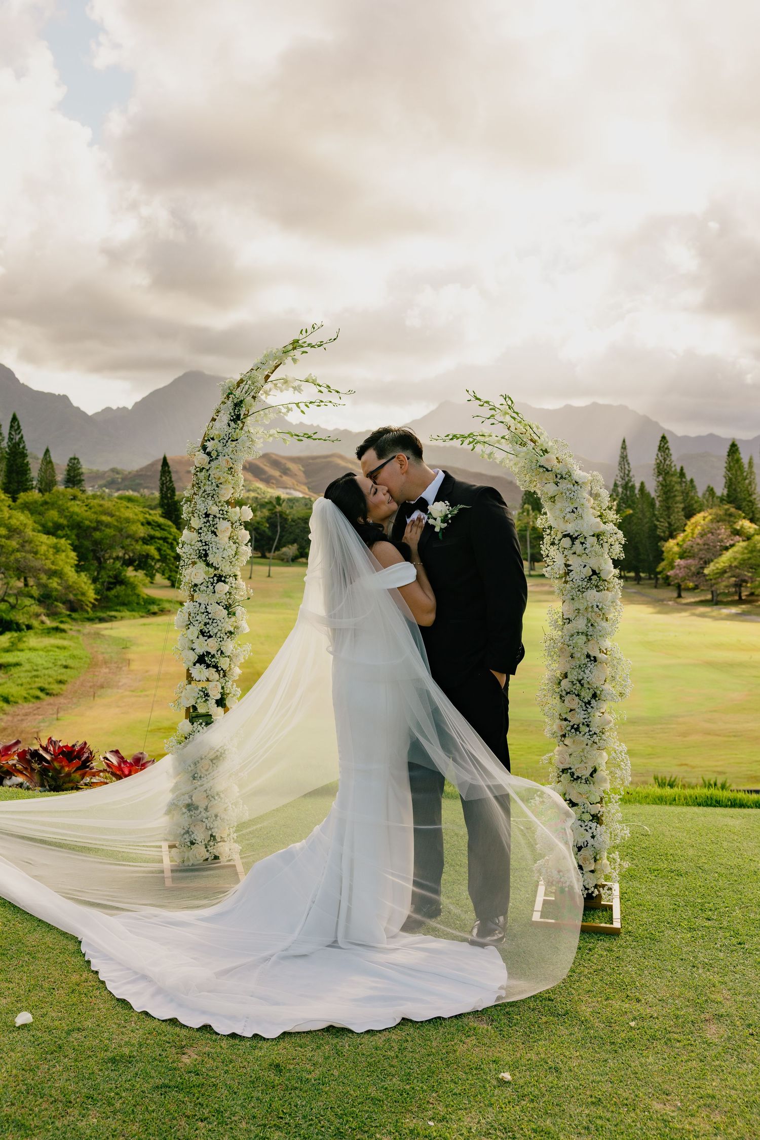 Bride and groom kissing under a floral arch with mountains and greenery in the background.