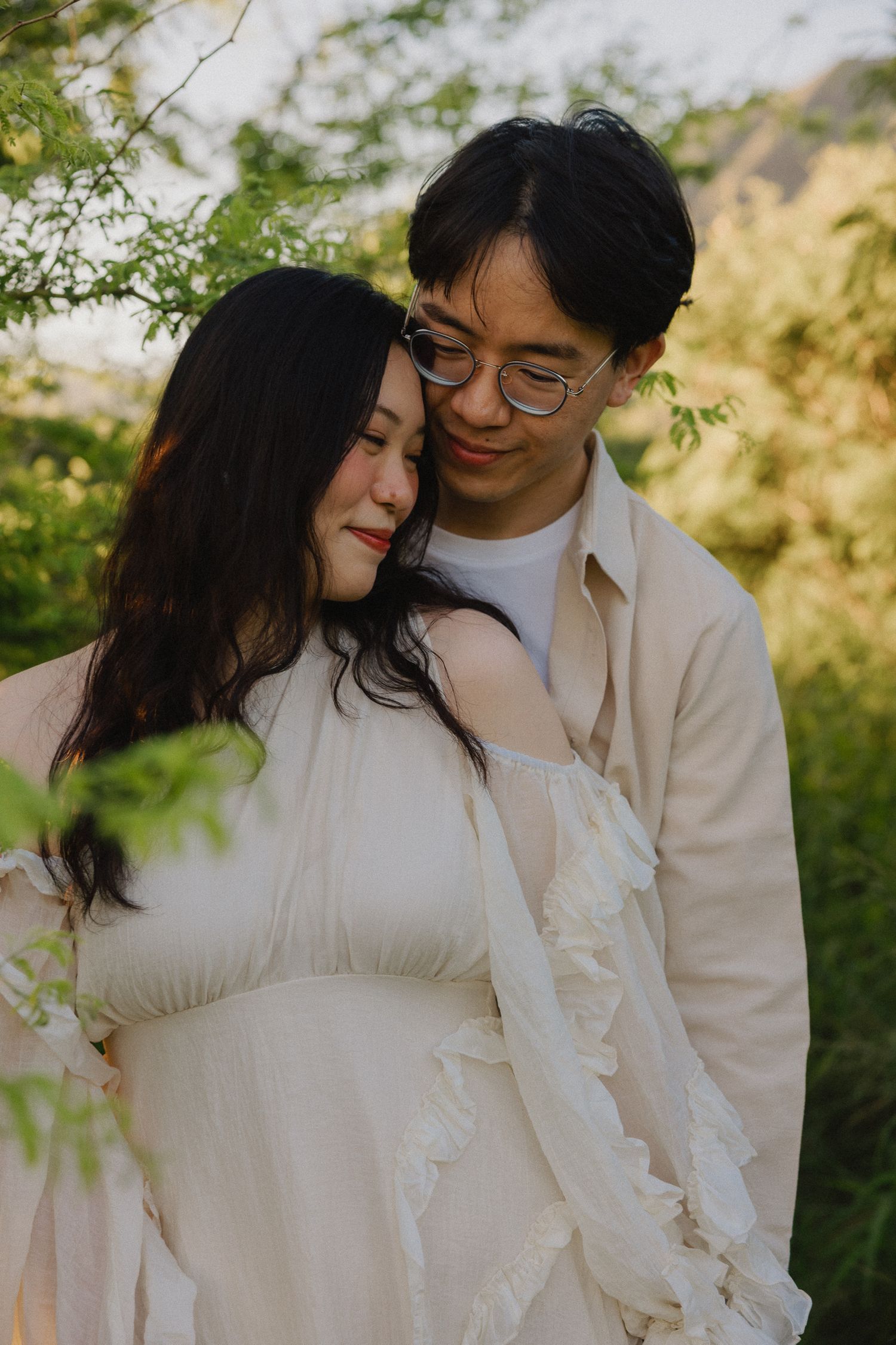 A couple shares a tender moment outdoors, woman in white ruffled maternity dress, man in beige shirt with glasses.