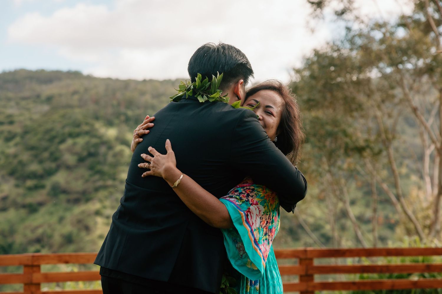Couple sharing a warm embrace outdoors, man wearing floral lei, woman in teal dress, lush green hills in background.