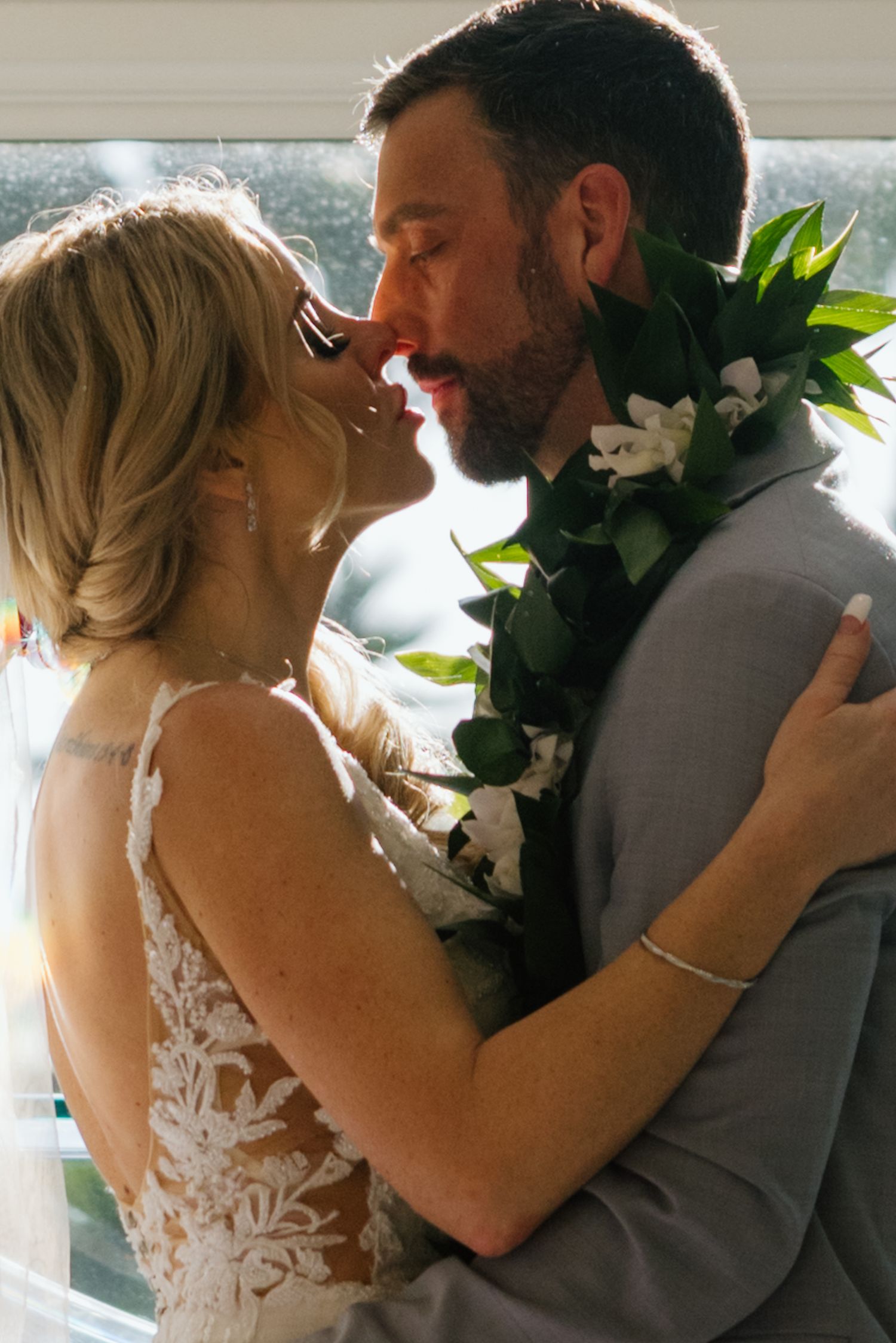 A bride and groom share a romantic moment, the groom wearing a floral lei, in warm backlit wedding photography.