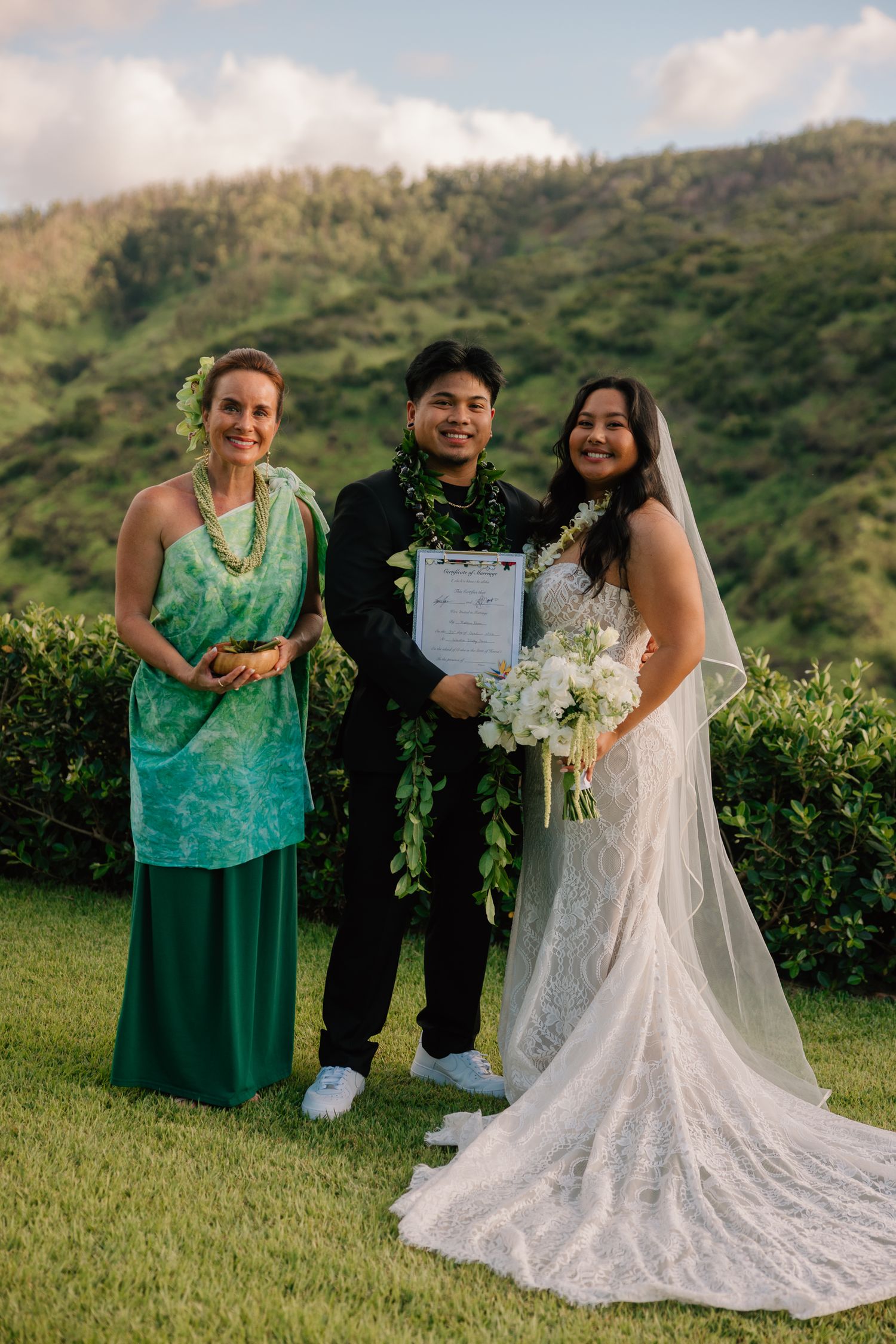 Bride and groom holding marriage certificate with officiant at outdoor Hawaii wedding ceremony with lush green mountains.