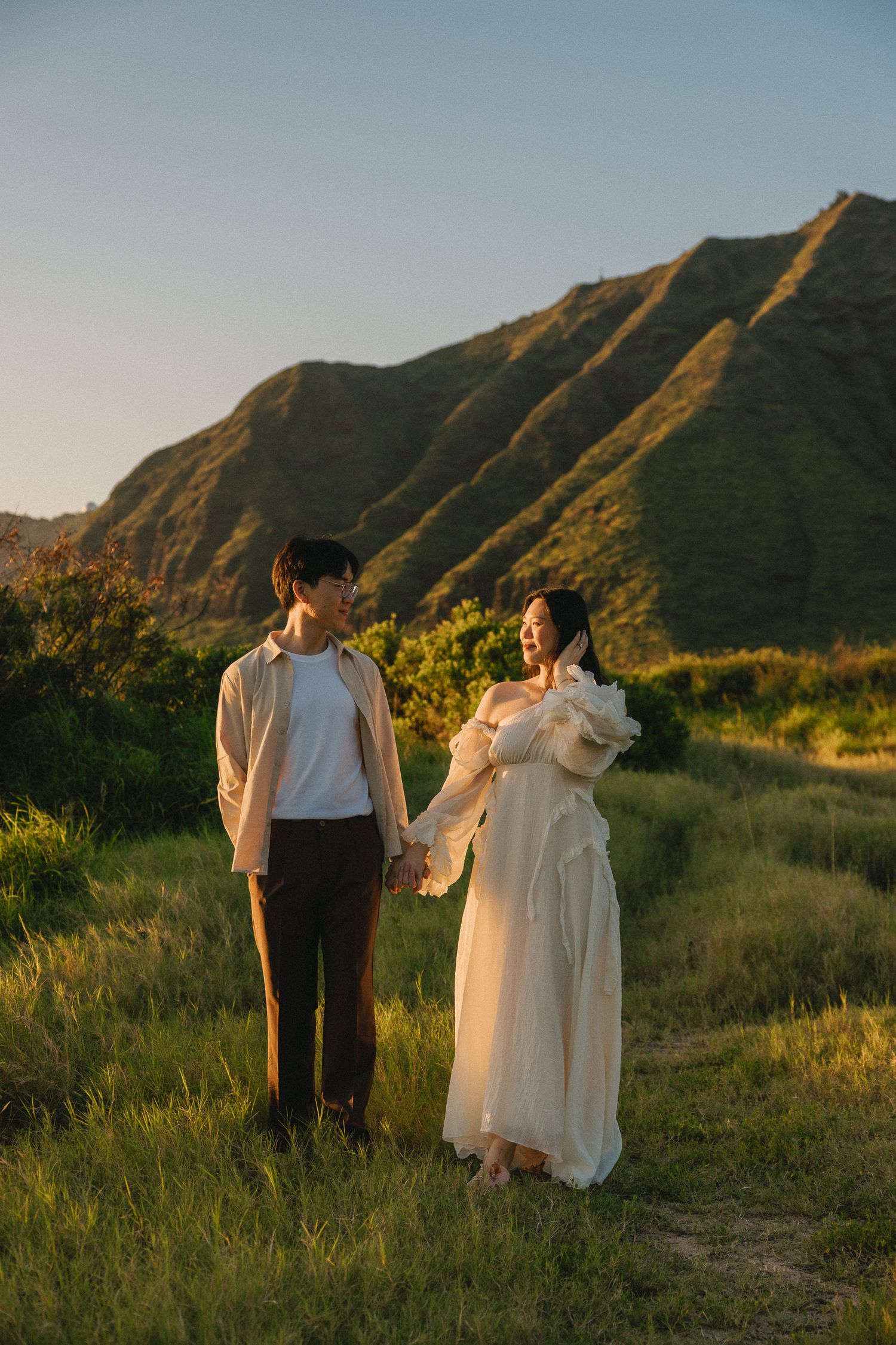 Couple holding hands in a grassy field with scenic green mountains at golden hour sunset.