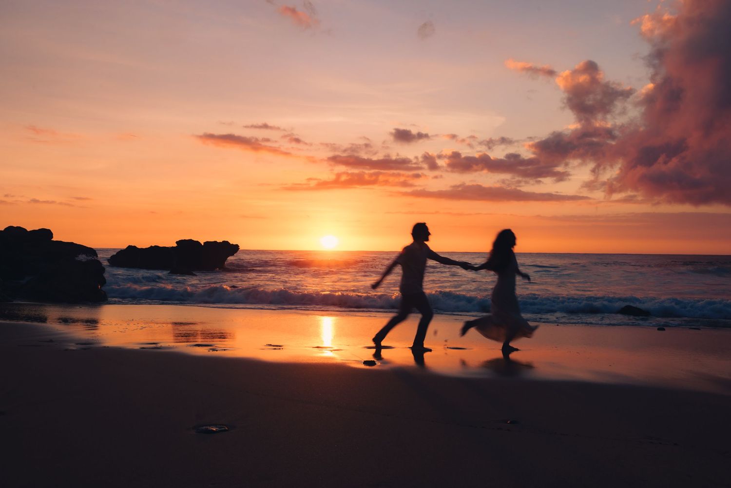 Silhouette of a couple holding hands running on a beach at sunset with vibrant orange and pink sky.