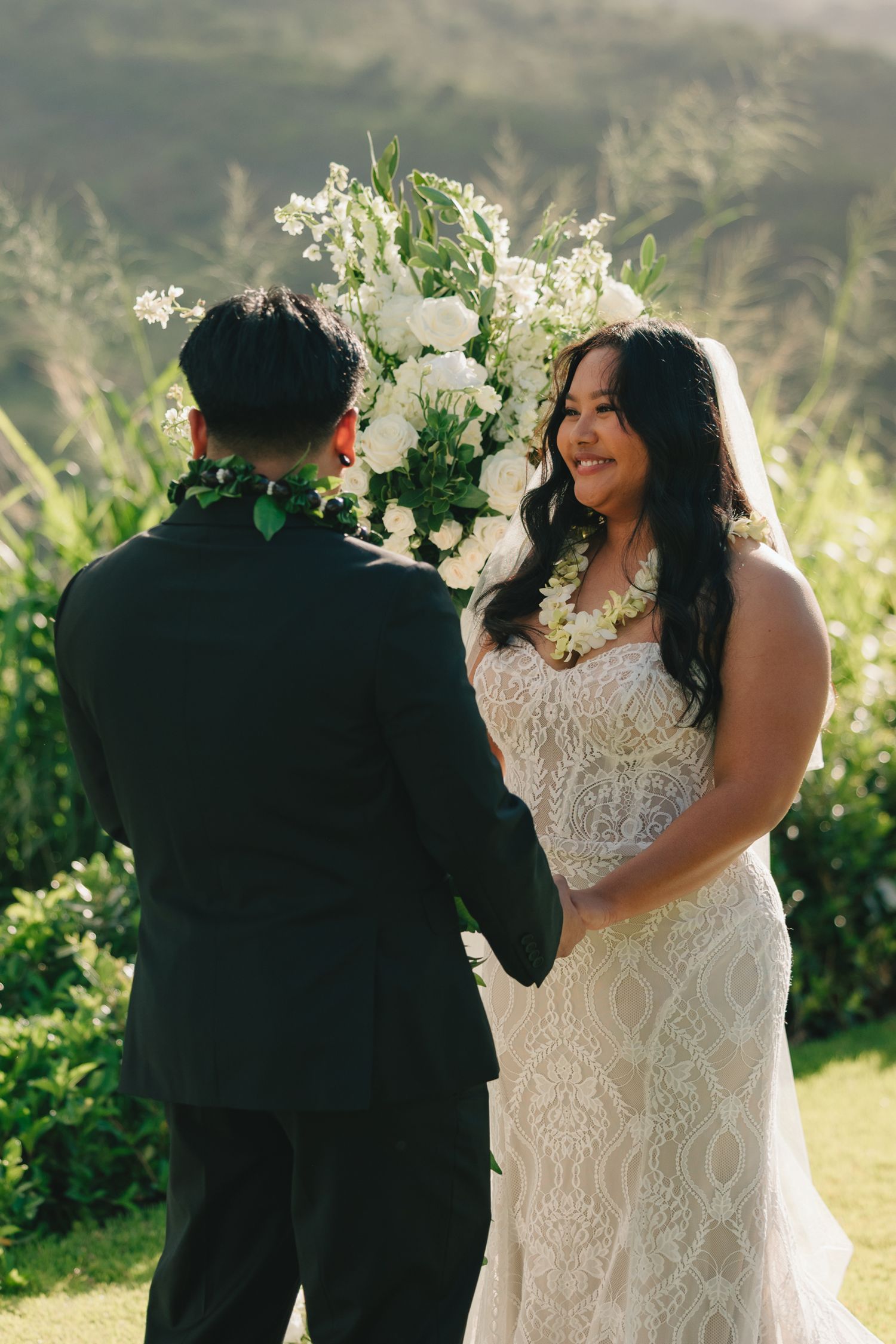 Bride in lace gown holds hands with groom during outdoor Hawaii wedding ceremony with white floral arch backdrop.
