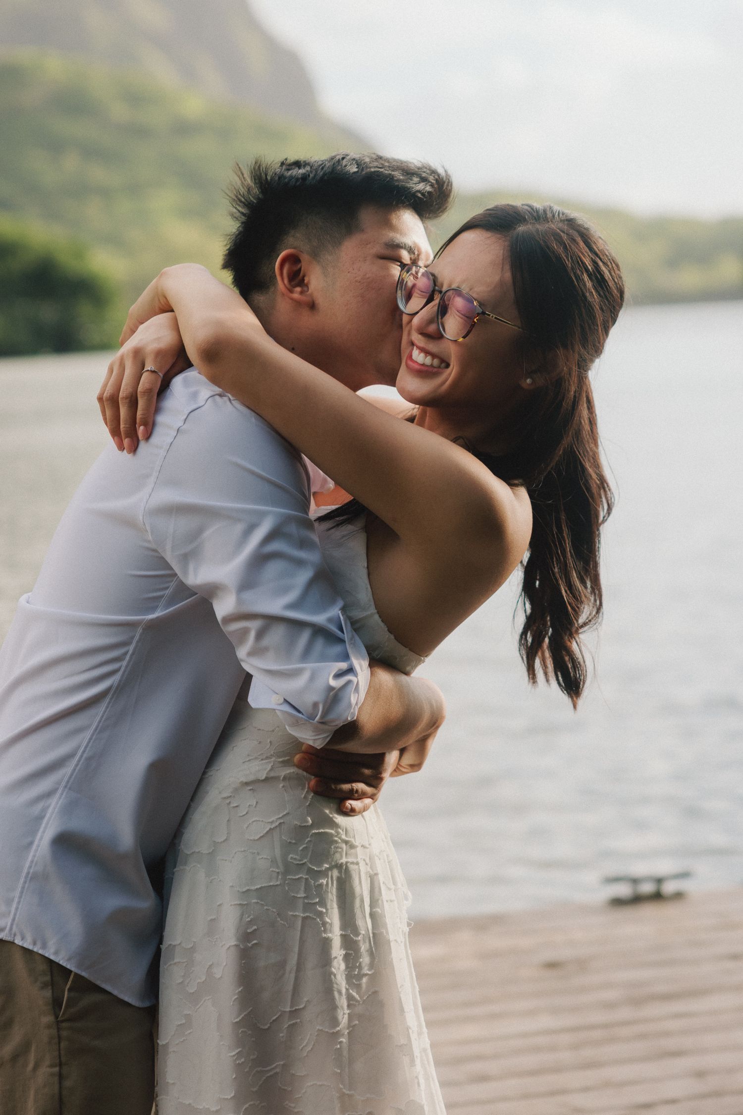 A joyful couple embraces and laughs on a wooden dock with a scenic lake and green mountains behind them.