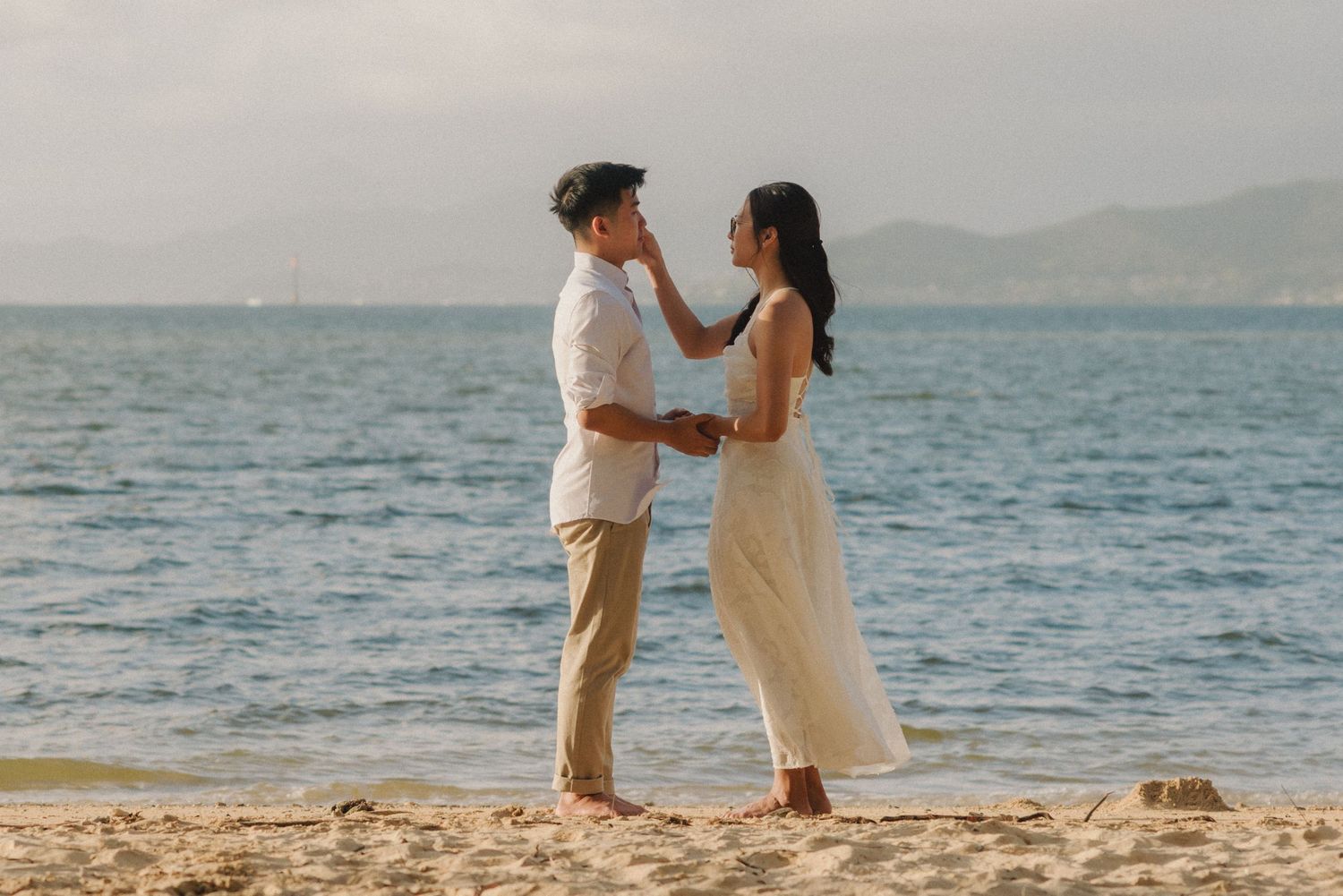 Romantic couple holding hands on a sandy beach with ocean and mountains in the background.