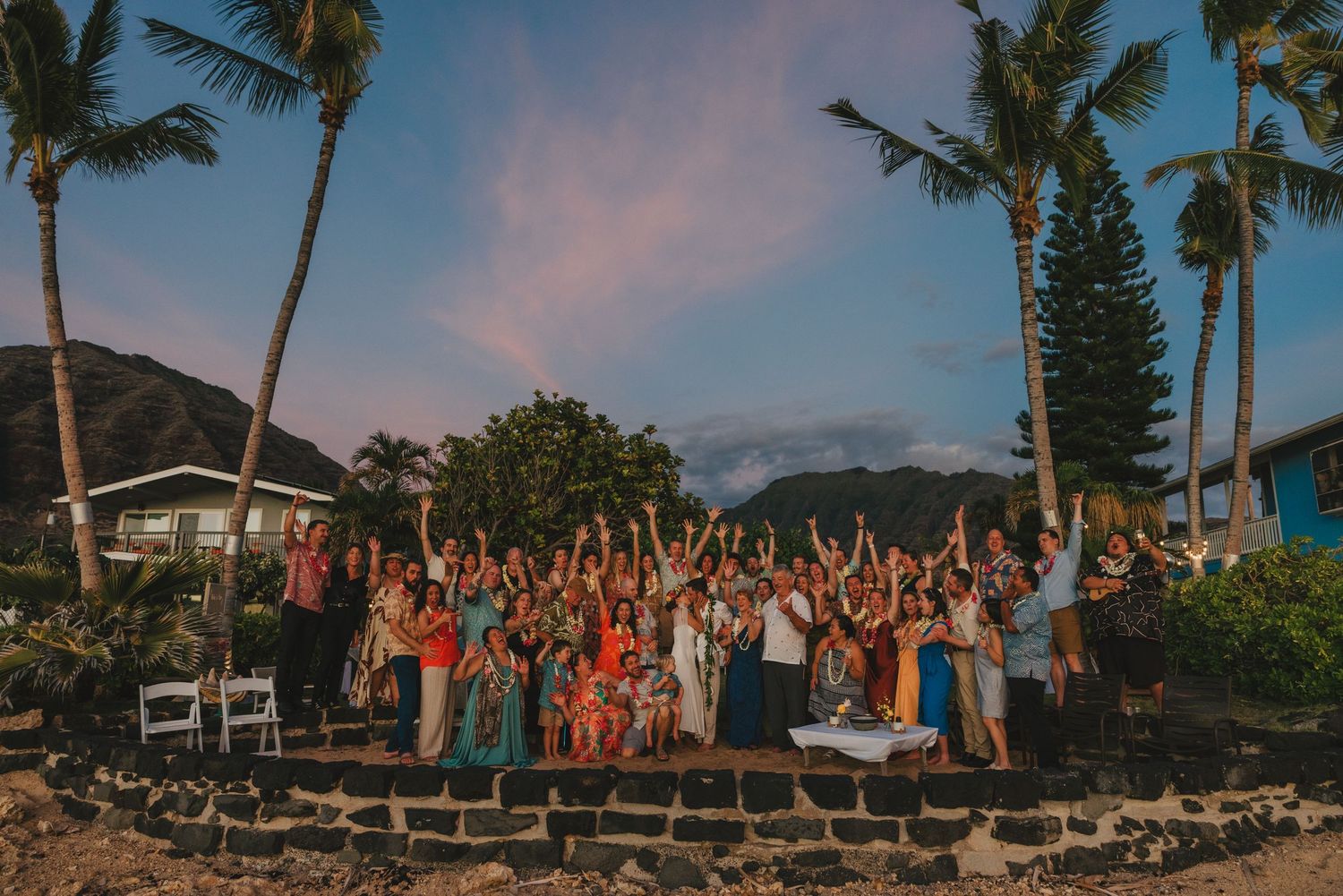 Large wedding party cheers under vibrant pink sunset sky surrounded by tall palm trees.