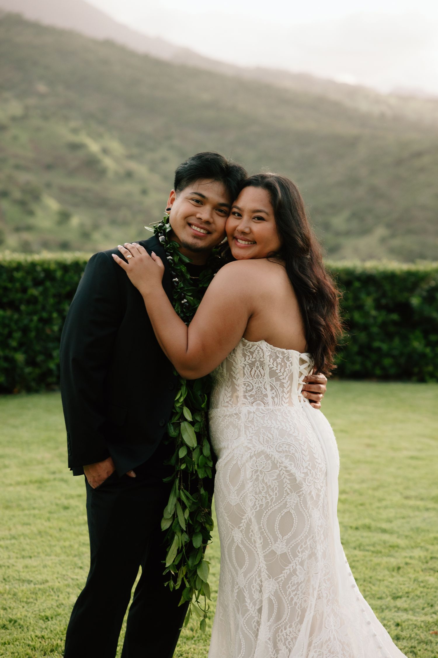 Bride in lace gown and groom in black suit with floral lei embrace outdoors with green hills backdrop.