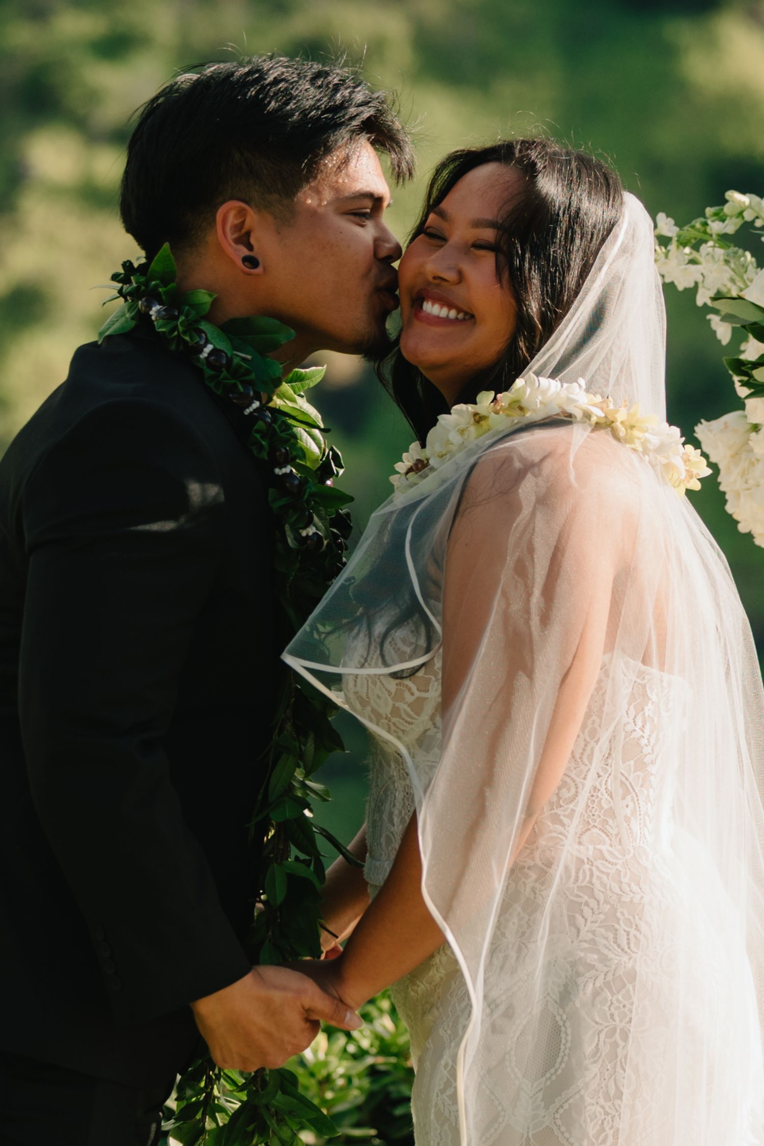 Groom kisses bride on cheek as she smiles, both wearing floral leis in a lush green outdoor wedding setting.