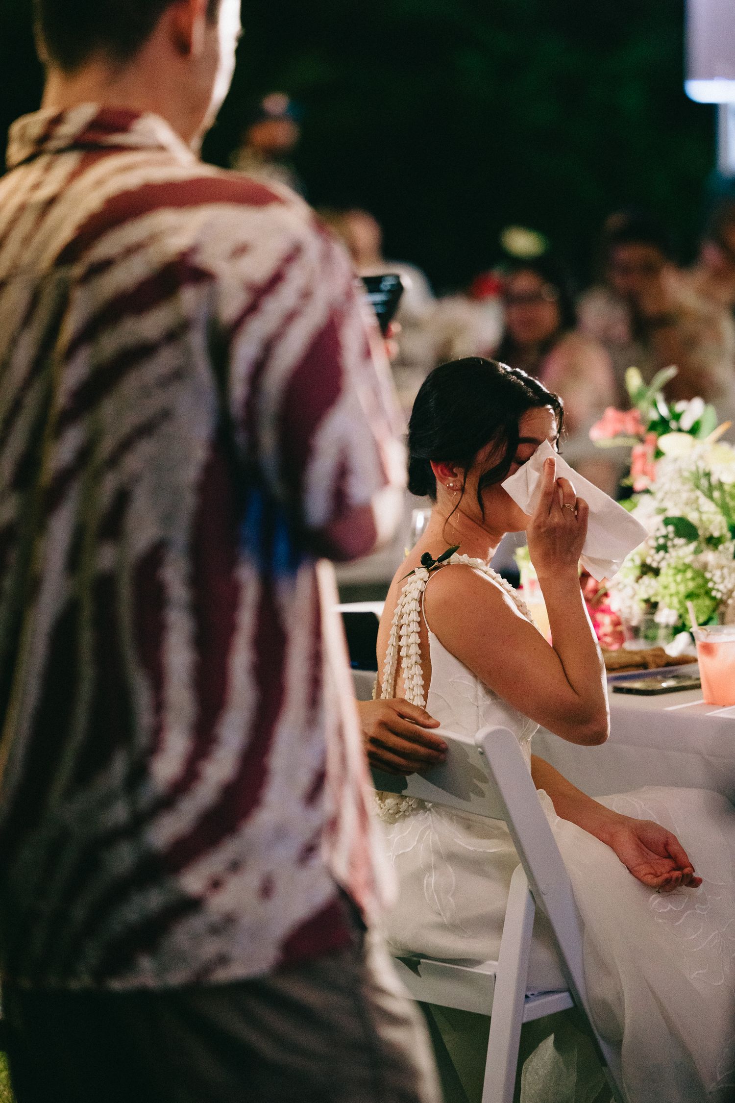 Emotional bride wiping tears with tissue at outdoor wedding reception dinner table.