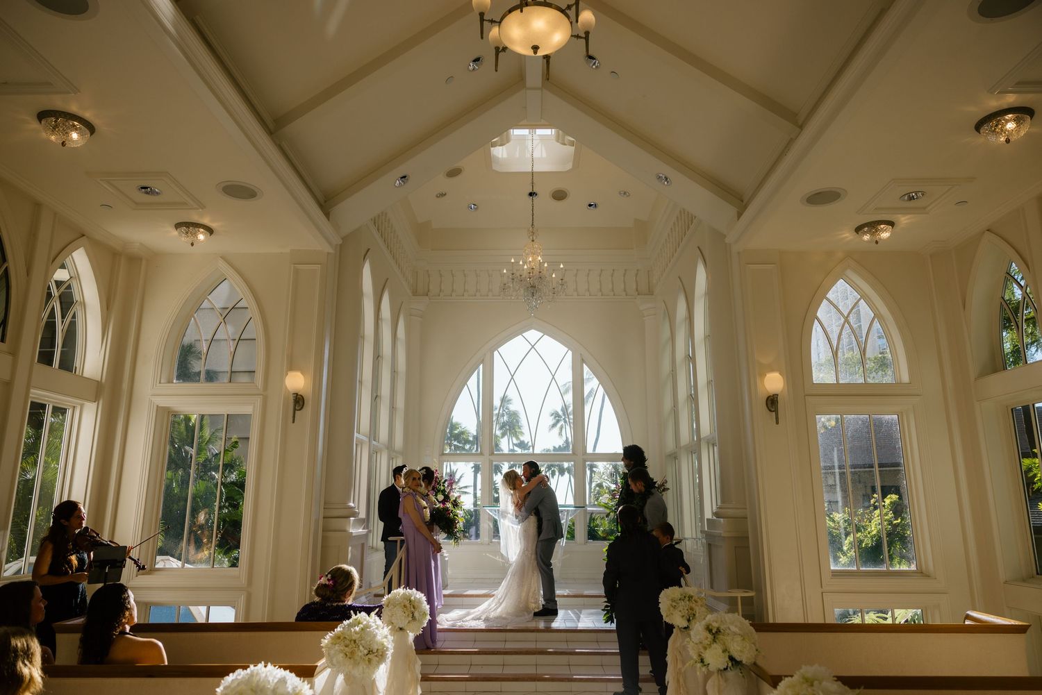 Elegant wedding ceremony in a white chapel with arched windows, couple kissing at altar with guests seated in pews.