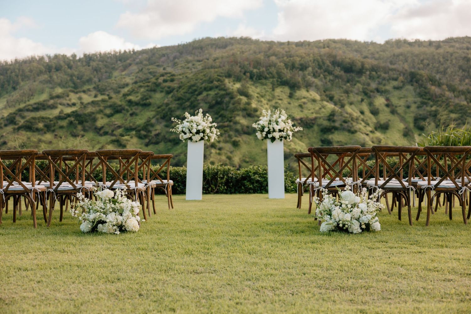 Outdoor wedding ceremony setup with white floral arrangements on pedestals, wooden cross-back chairs, and lush green mountain backdrop.