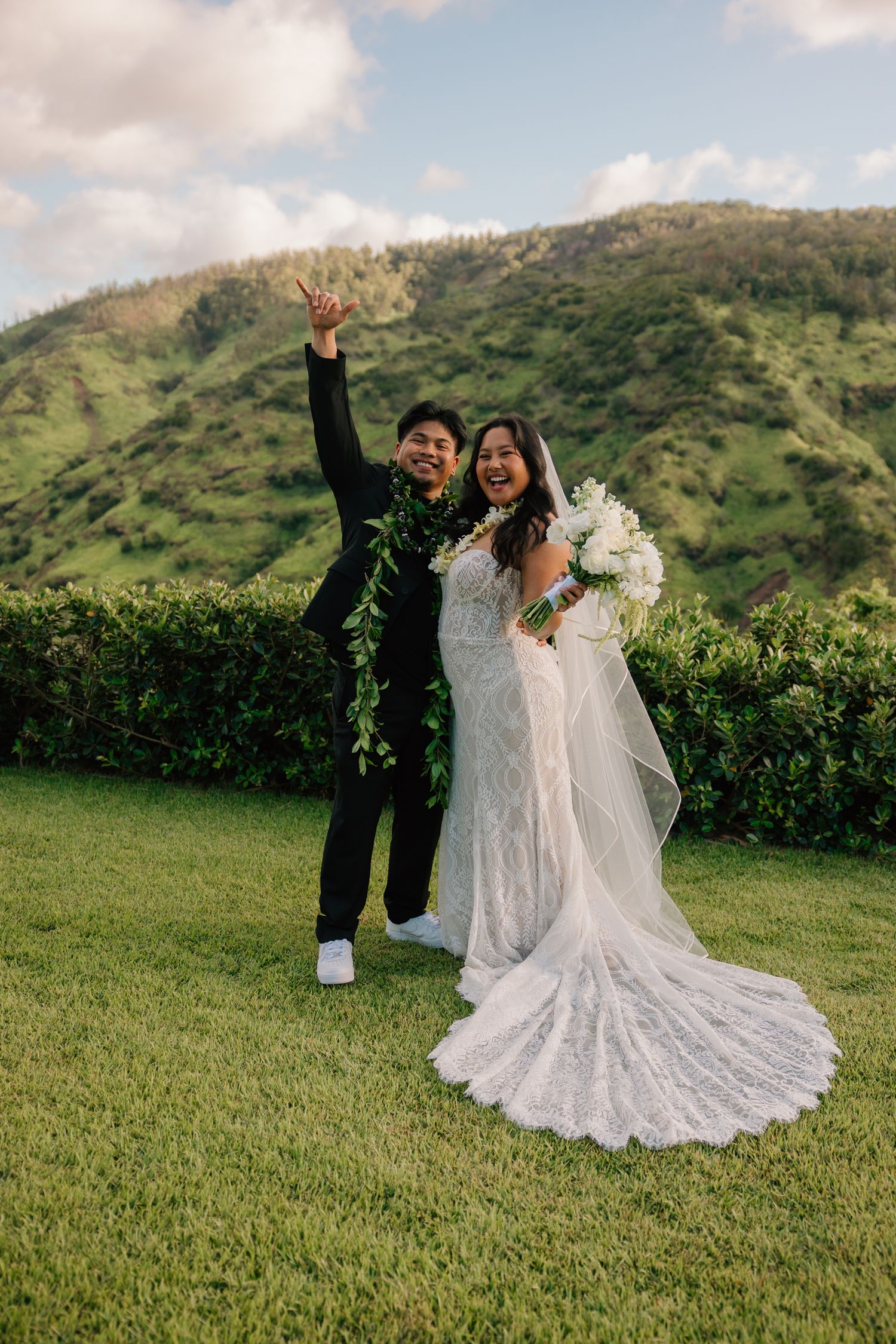 Joyful newlyweds celebrate on green lawn with lush Hawaiian mountains, groom in lei, bride in lace gown holding white bouquet.