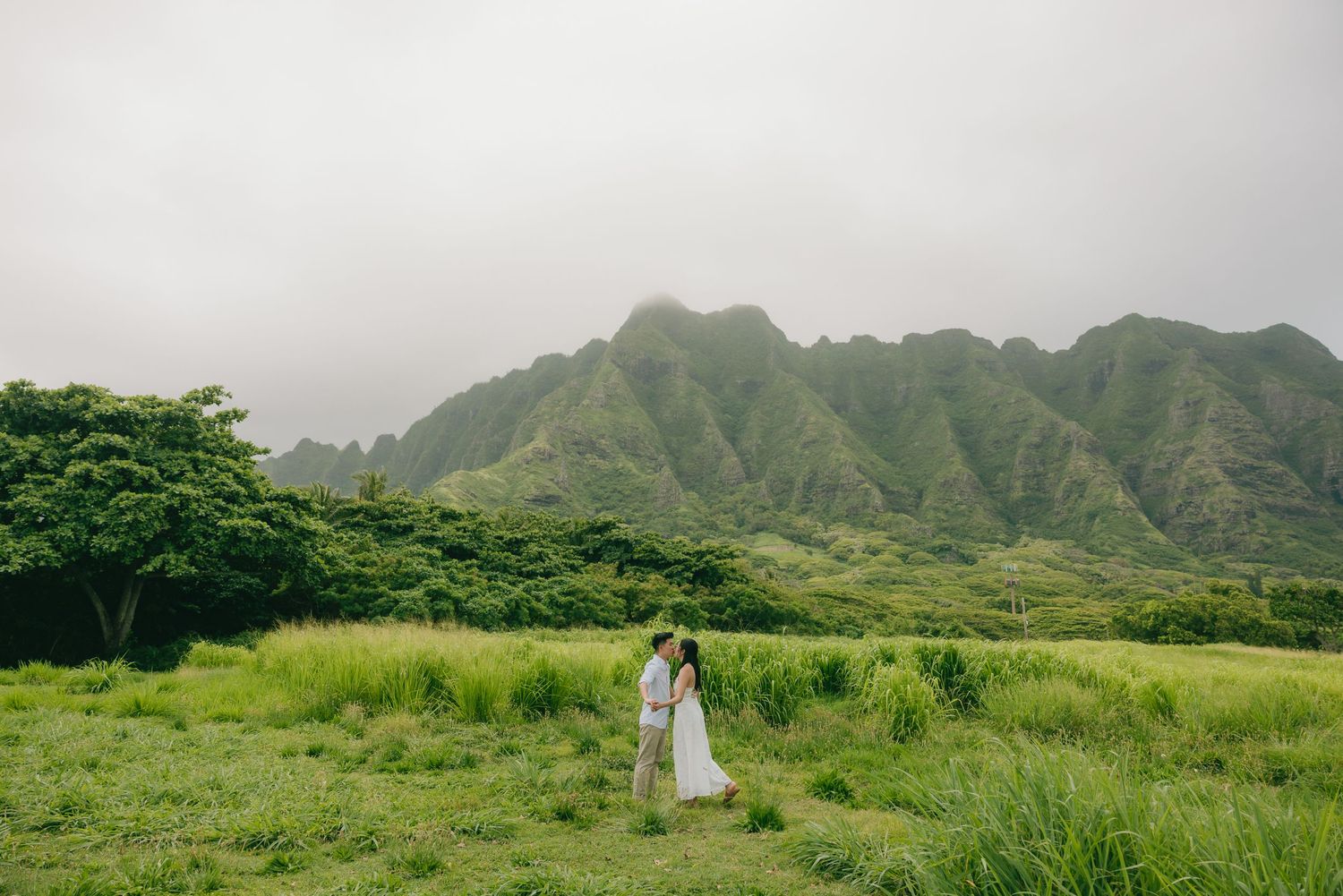 Couple embraces in lush green meadow with dramatic misty Ko'olau Mountains backdrop in Hawaii.