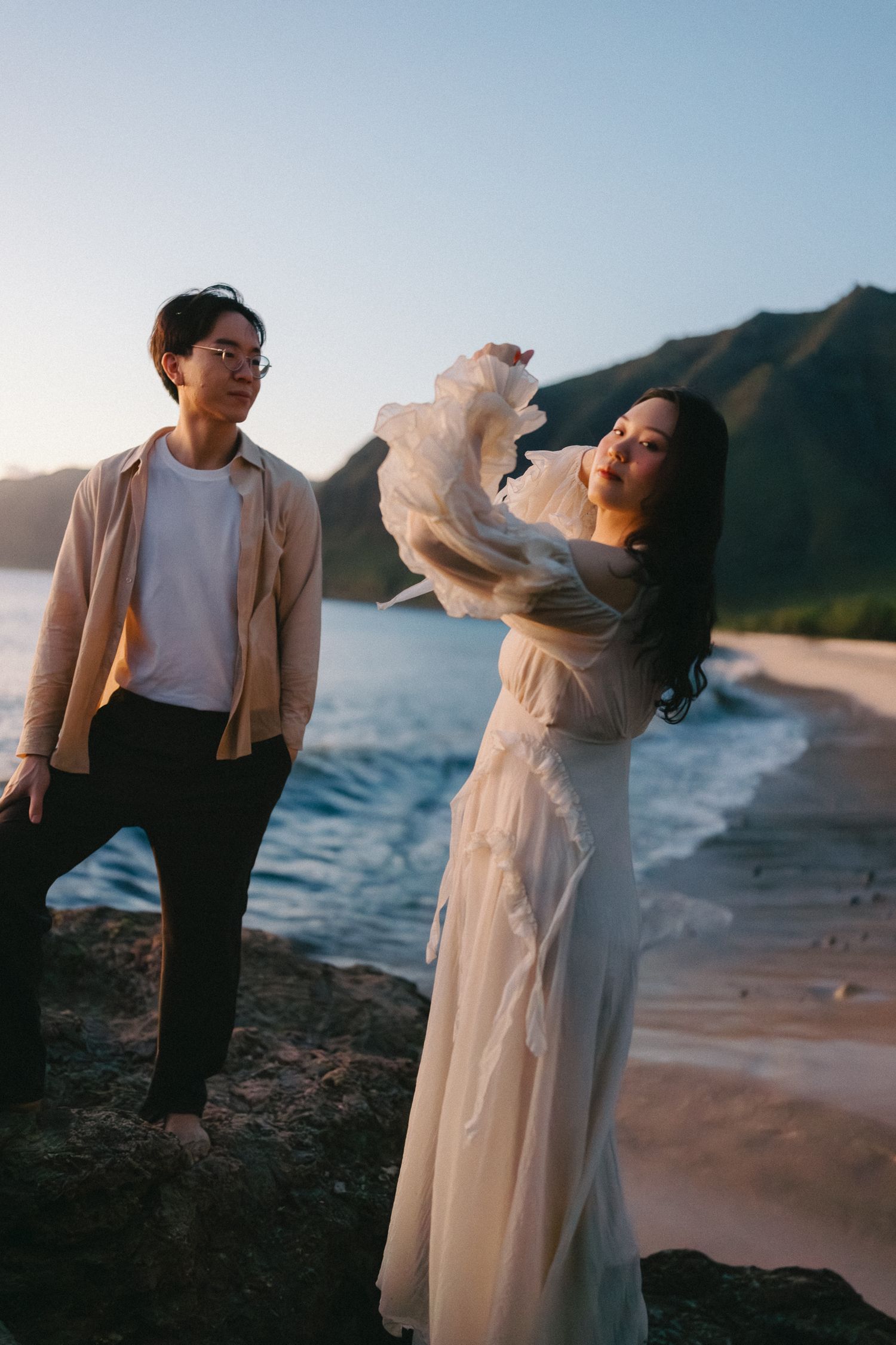 Couple in romantic beach photoshoot at sunset, woman in flowing white dress, man in beige jacket, with ocean and mountains.