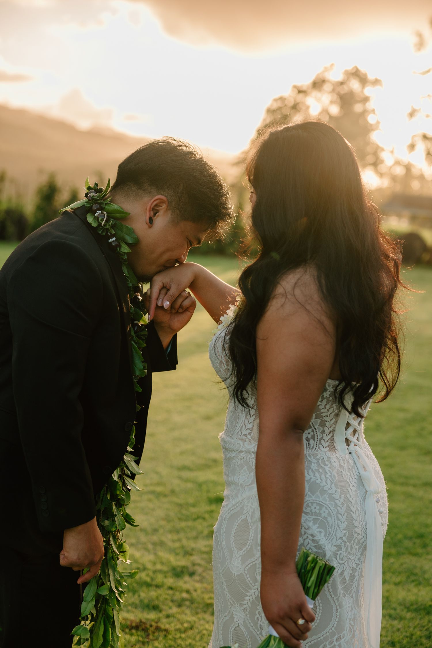 Groom in black suit kisses bride's hand at golden sunset, both wearing Hawaiian leis, bride in lace wedding gown outdoors.