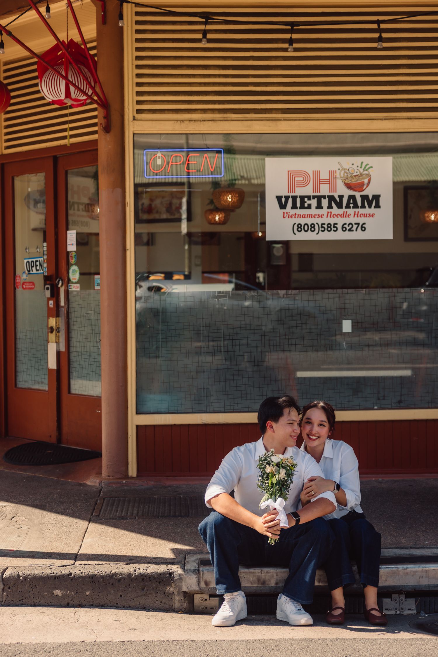 Couple sitting on steps outside Pho Vietnam Vietnamese Noodle House, sharing a romantic moment with flowers.