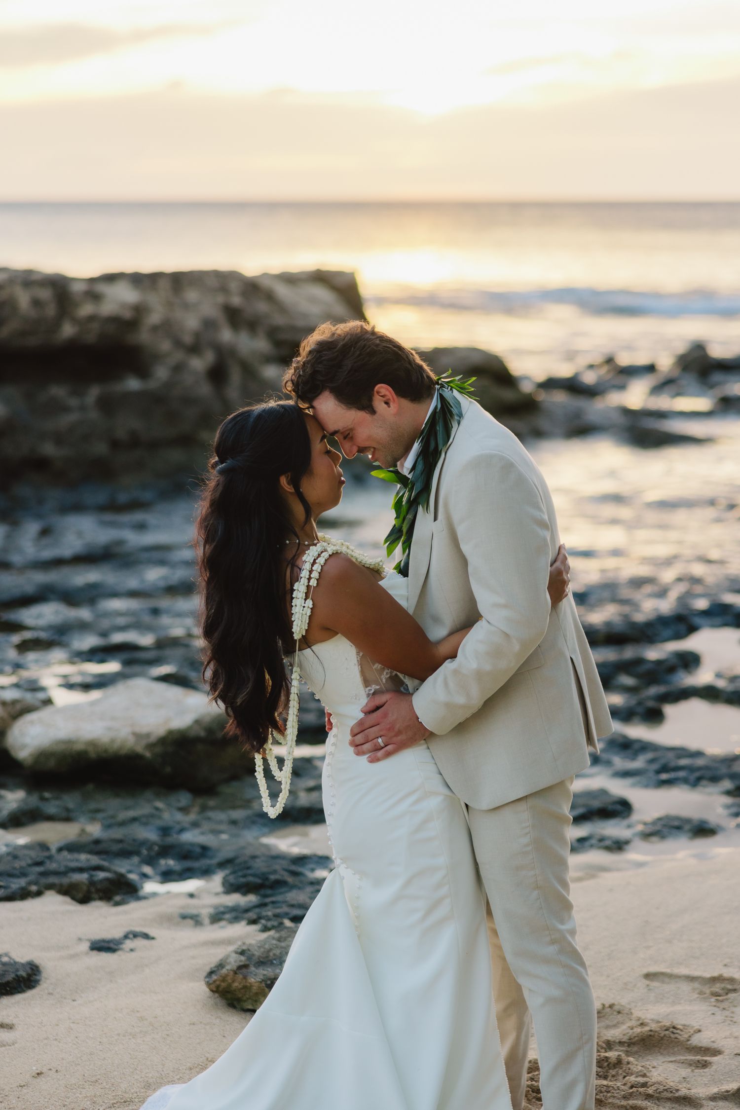 Bride and groom share a romantic beach moment at sunset, she in a white gown, he in a tan suit with floral lei.