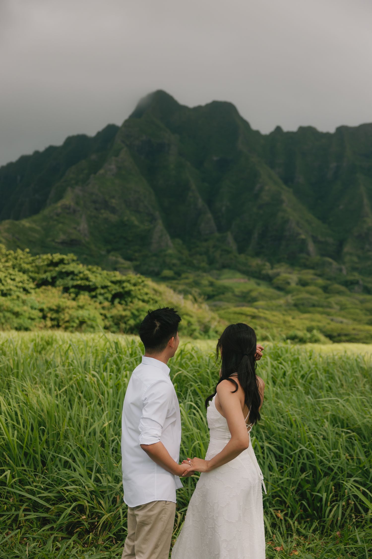 Couple holding hands facing lush green mountains in Hawaii, wearing white outfits in a romantic outdoor engagement photo.