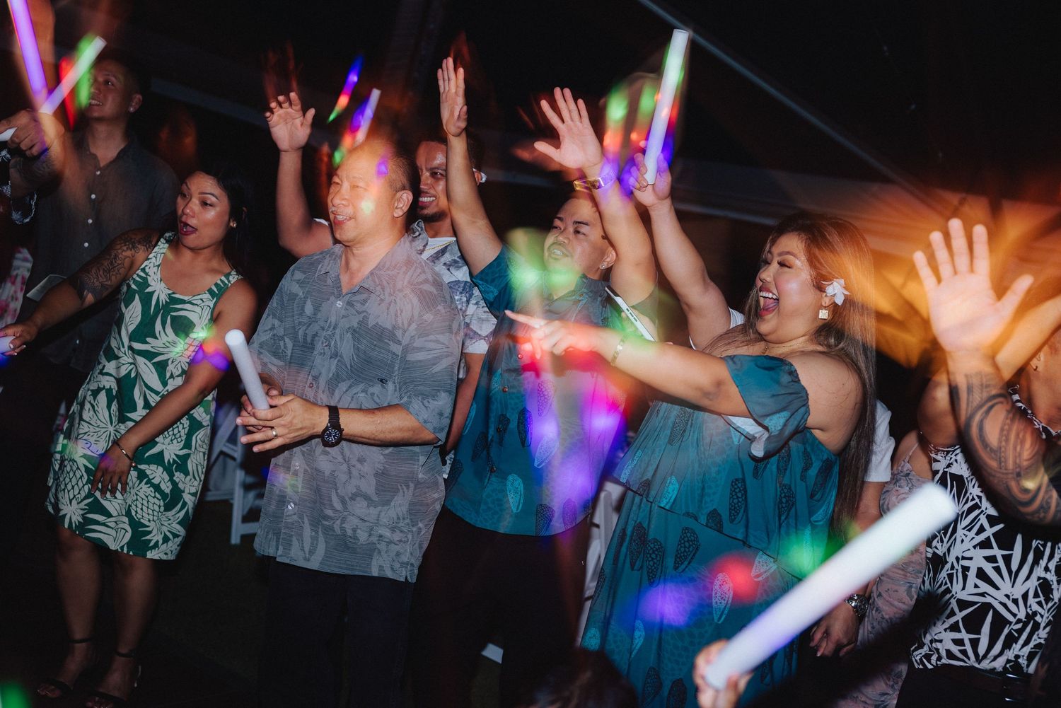 Guests dancing and celebrating at a party, holding colorful glowing light sticks under dark night sky.