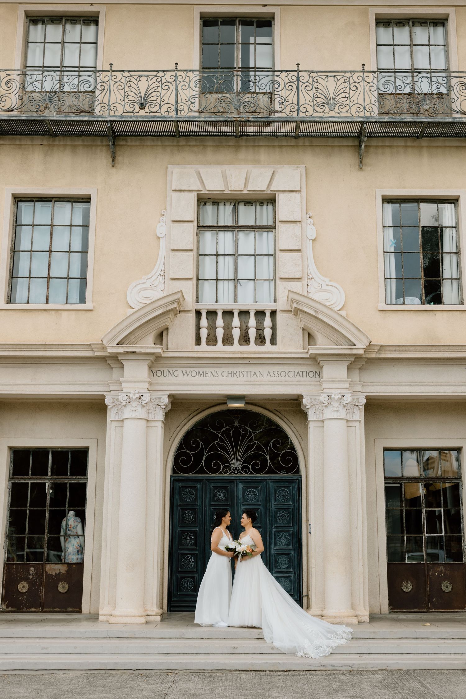 Two brides in white gowns standing before ornate doors of a historic YWCA building.