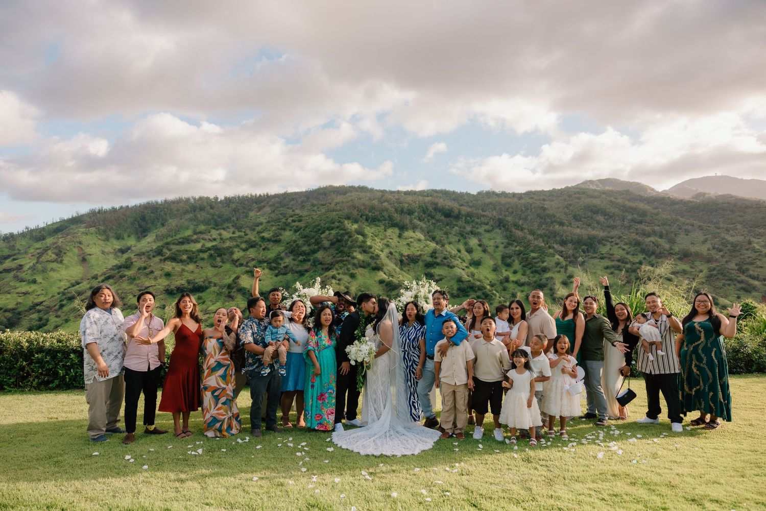 Wedding group photo with bride and groom surrounded by guests on green lawn with lush mountain backdrop in Hawaii.