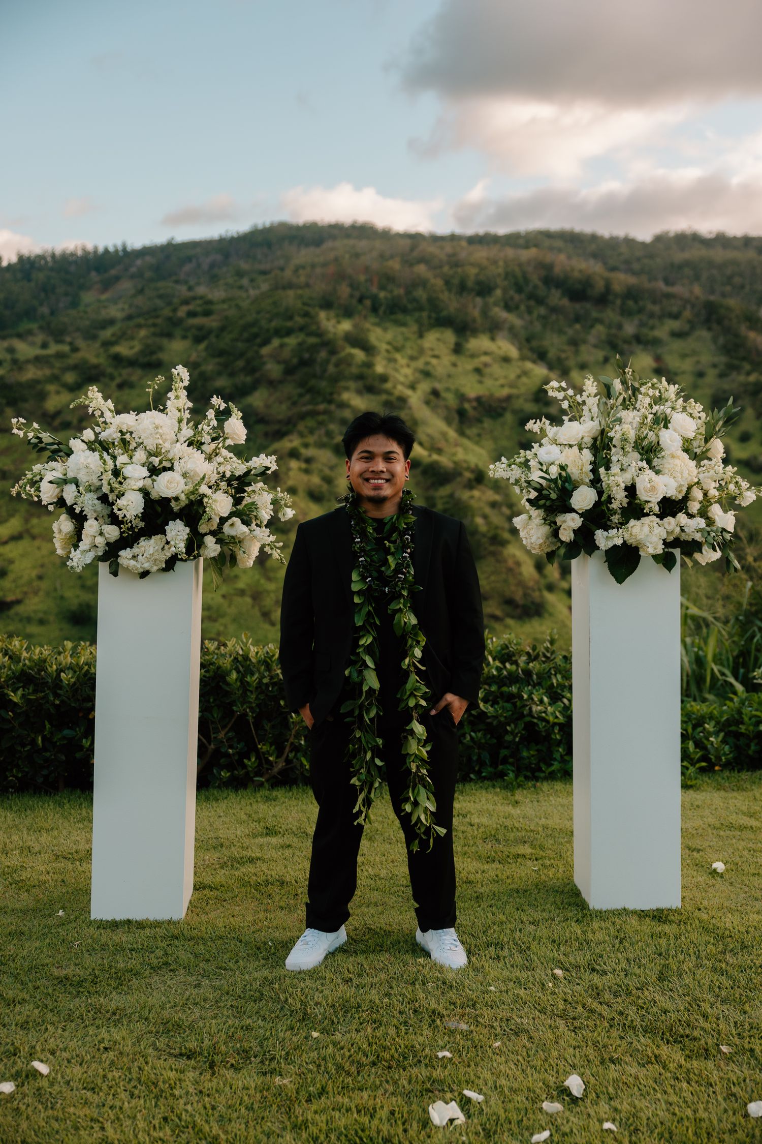 Man in black suit with green lei stands between white floral pedestals at outdoor wedding ceremony with green hills backdrop.