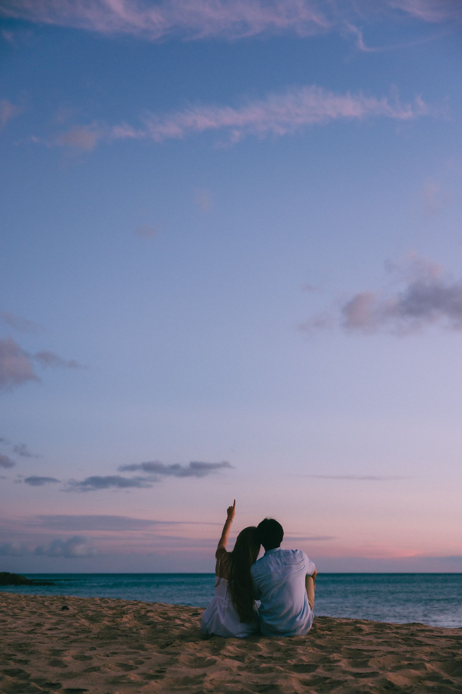 Couple sitting on sandy beach at sunset, one pointing at sky, with pastel pink and blue horizon over calm ocean.