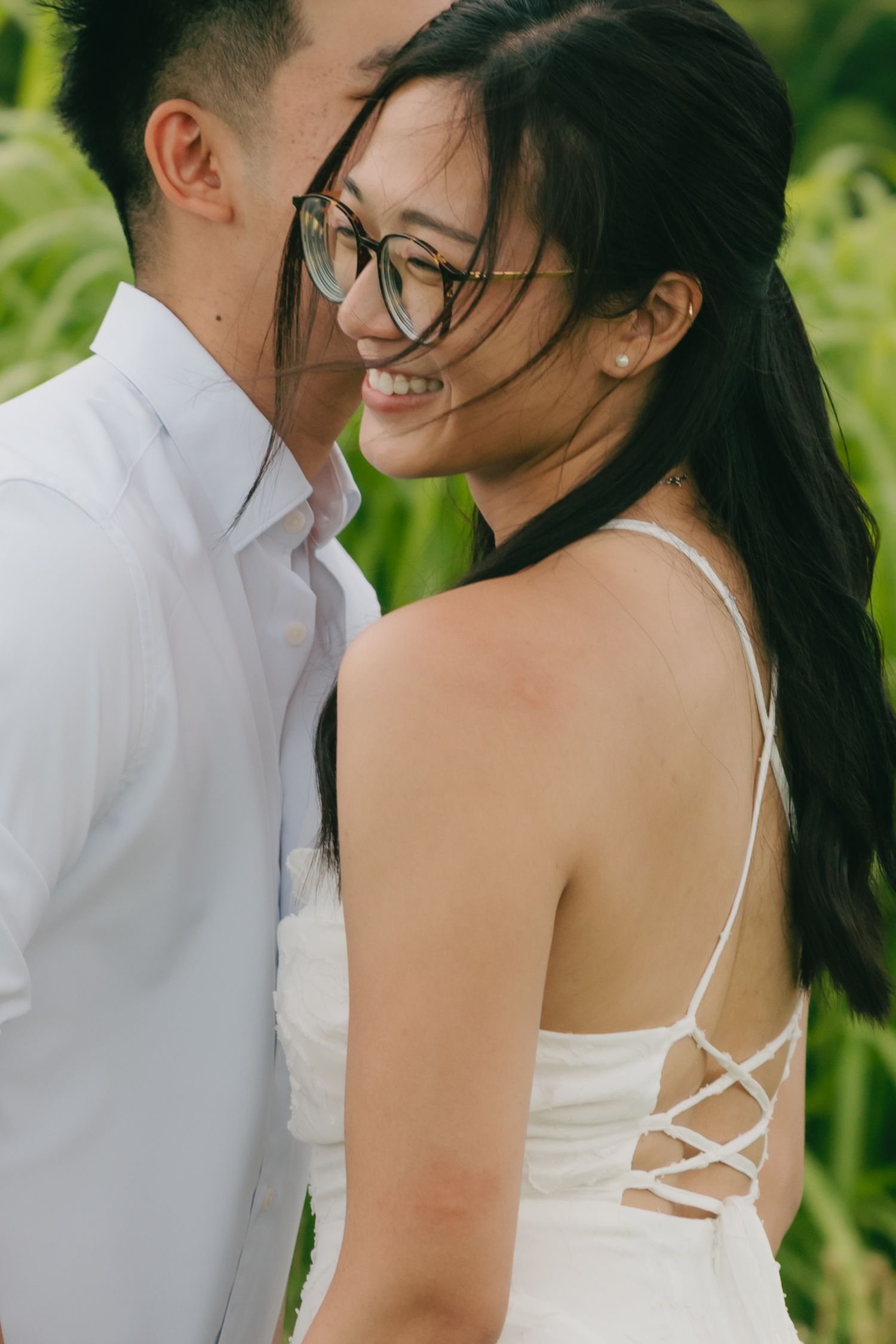 A smiling woman with glasses leans into her partner outdoors near green foliage.