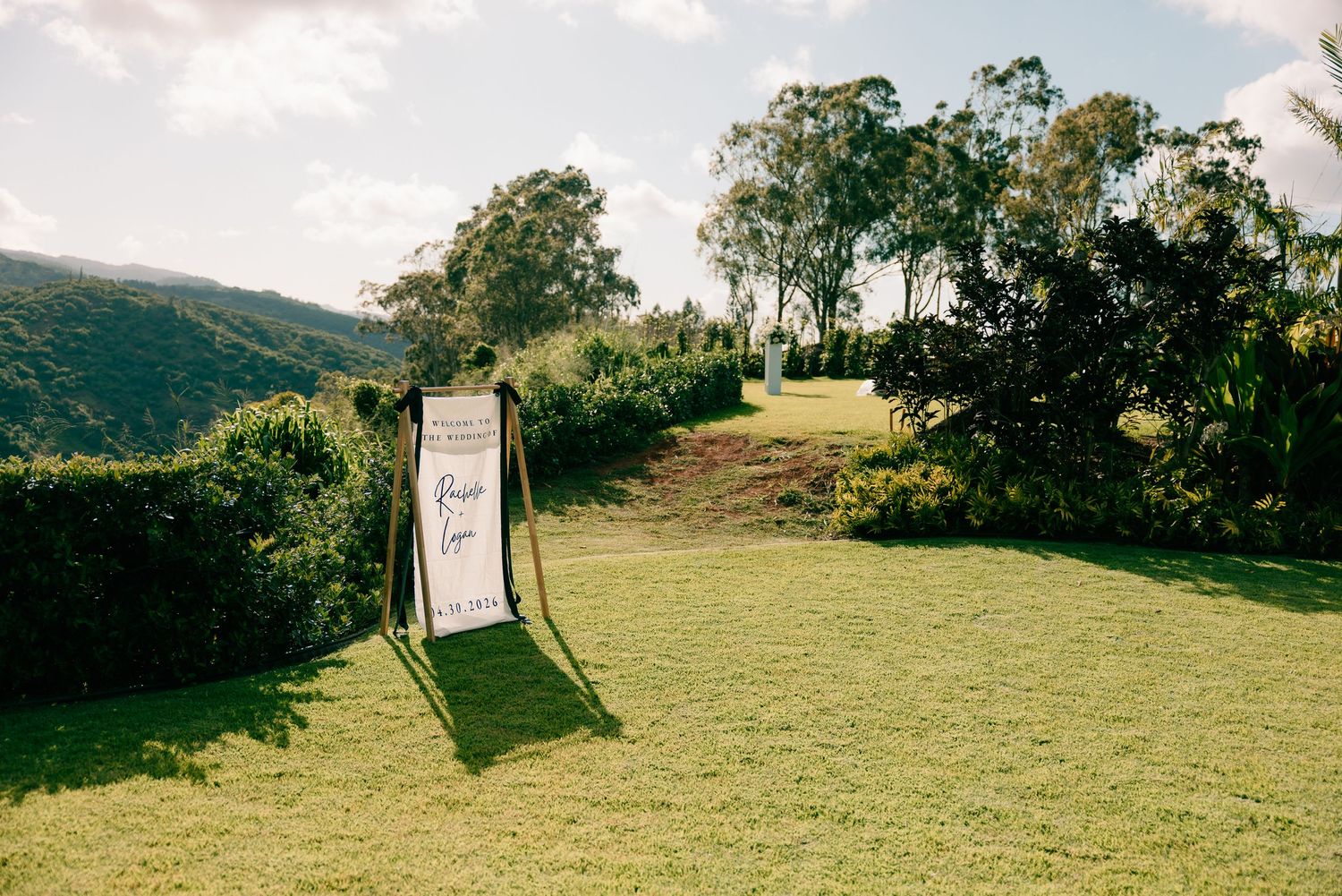 Outdoor wedding ceremony sign on lush green lawn with scenic mountain and tree backdrop on a sunny day.