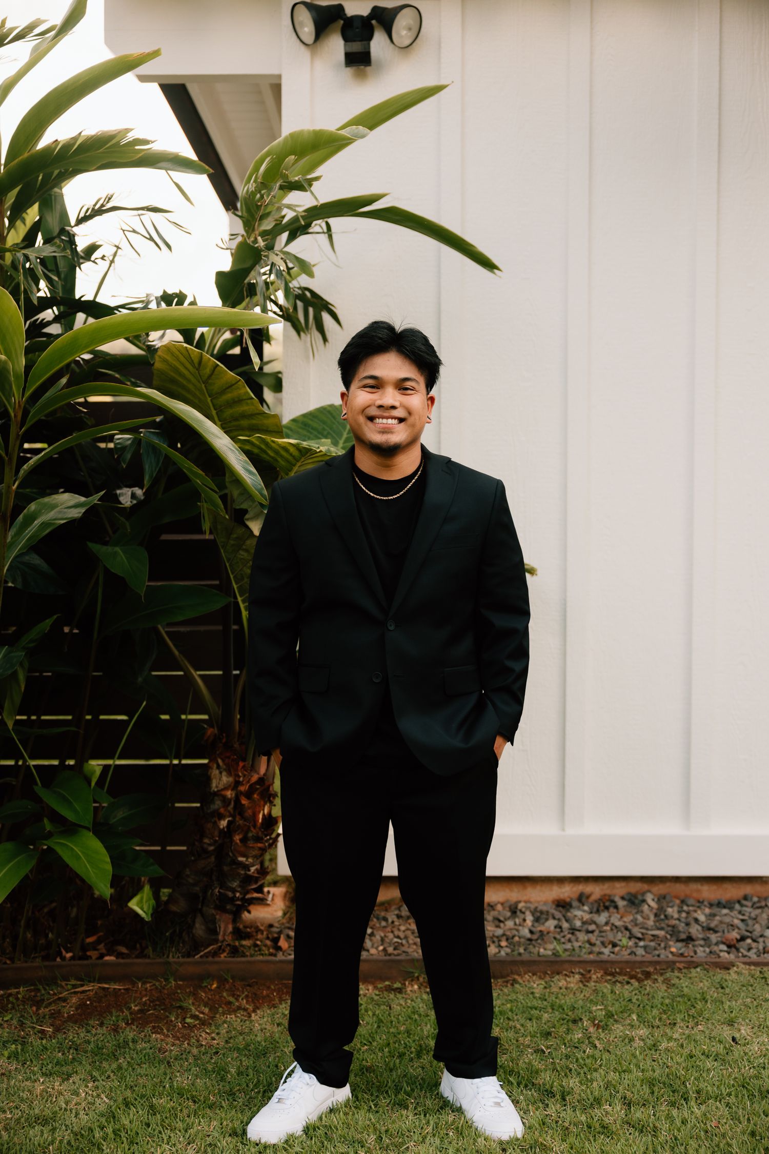 Young man smiling in black suit and white sneakers, standing outdoors near tropical plants by a white building.