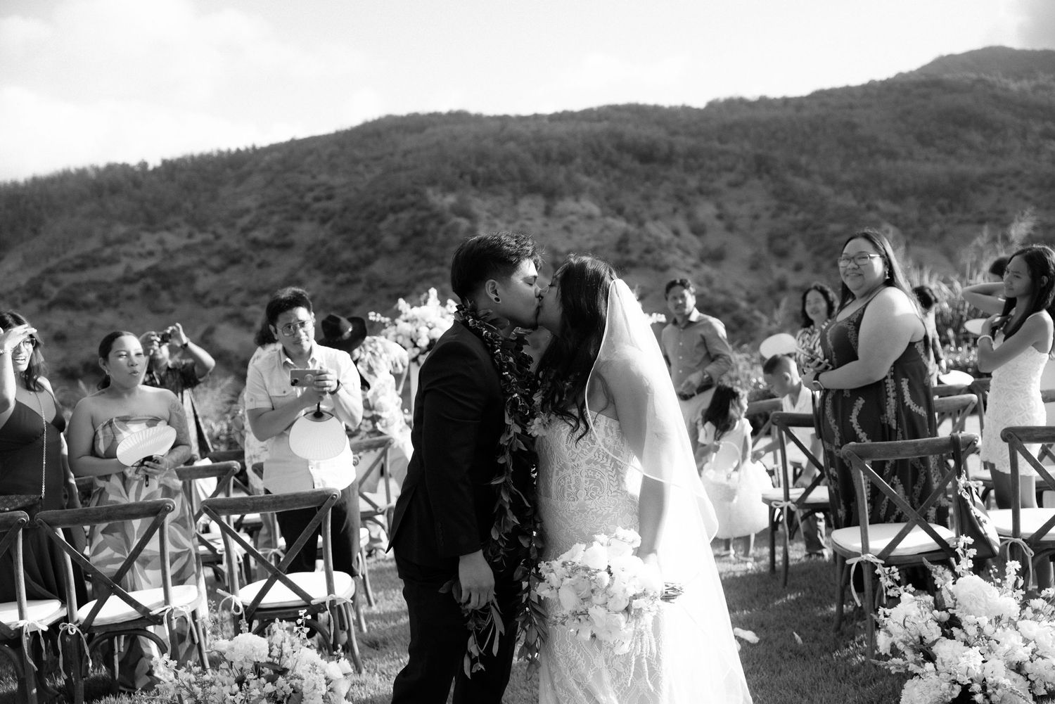 Black and white photo of a couple sharing their first kiss at an outdoor wedding ceremony with guests seated nearby.