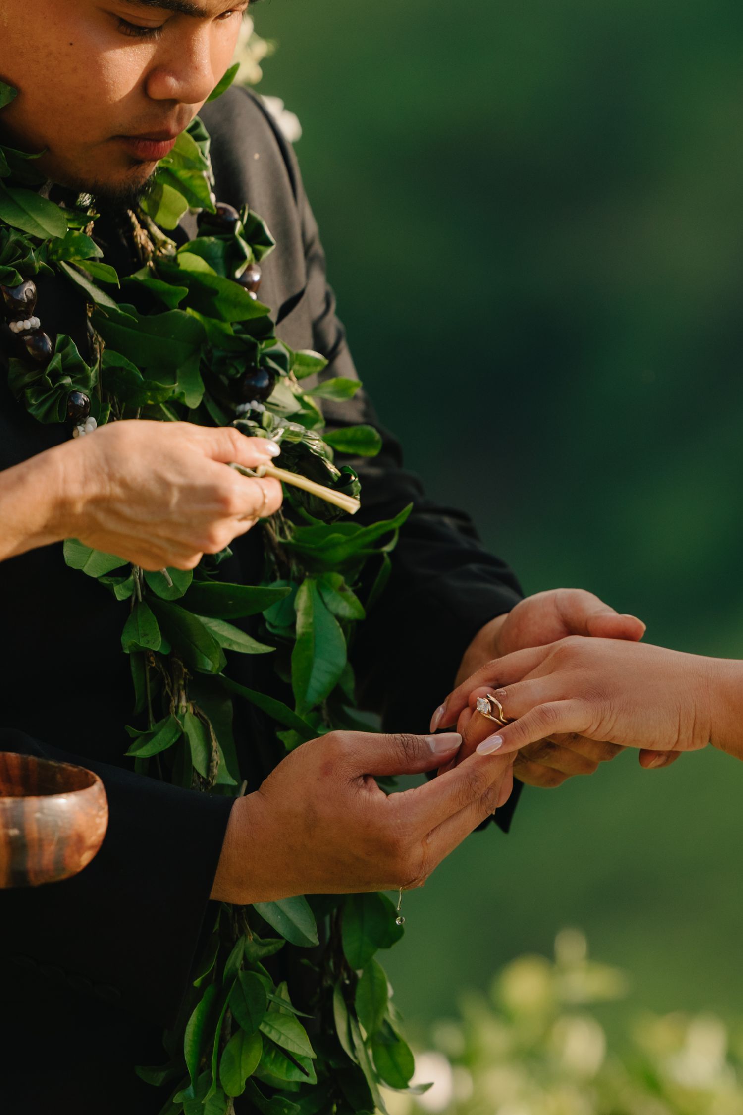 Hawaiian officiant wearing leaf lei performs ring ceremony outdoors in lush green setting.