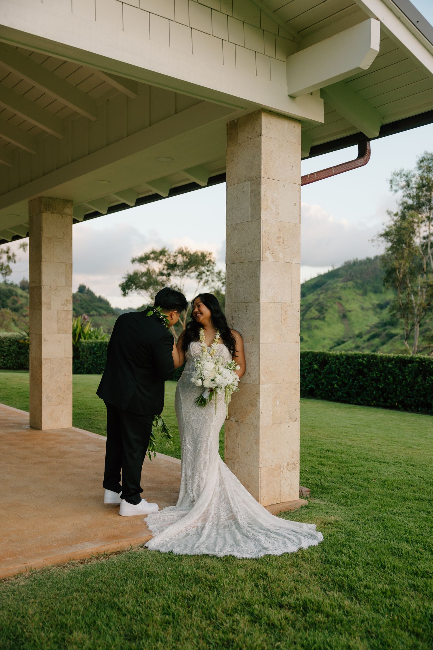 Bride in lace gown holds white bouquet as groom leans in tenderly under pavilion with lush green hills backdrop.
