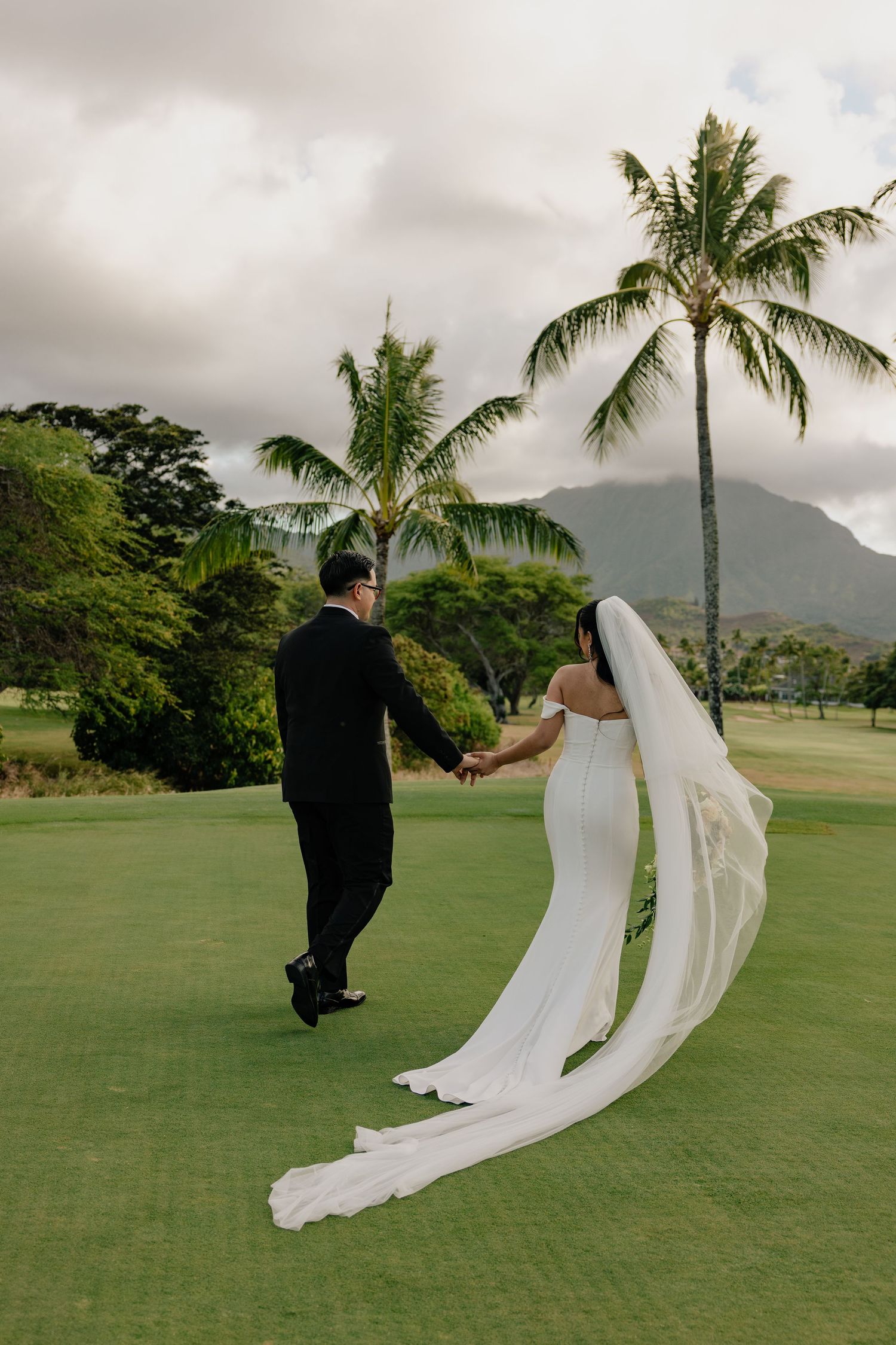 Bride and groom walking hand in hand on a golf course with palm trees and mountains behind.