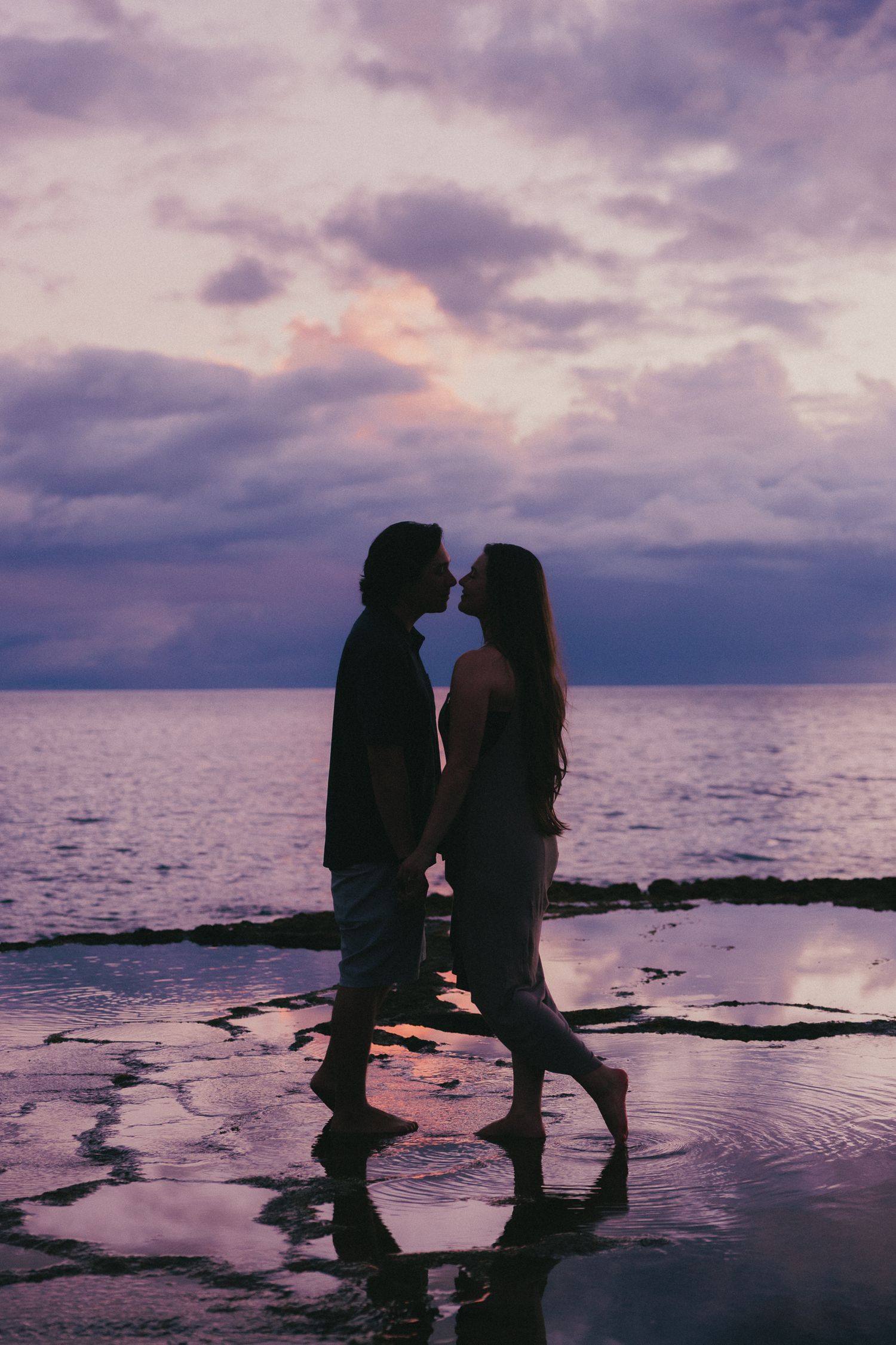Romantic couple silhouetted against a purple sunset sky, sharing a kiss on a reflective beach shoreline.