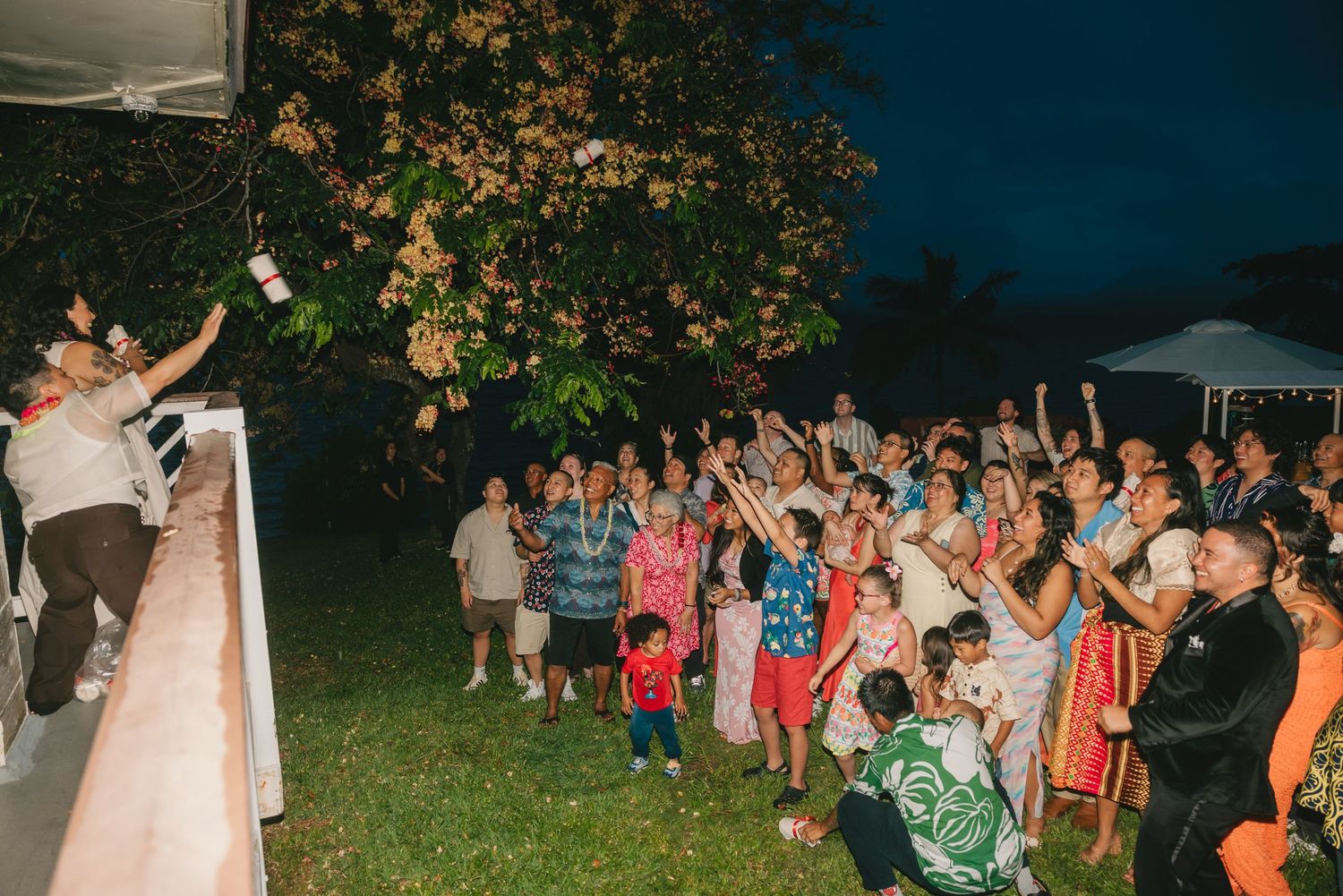 Guests cheer and raise hands at an outdoor evening wedding celebration under a tree with festive lighting.