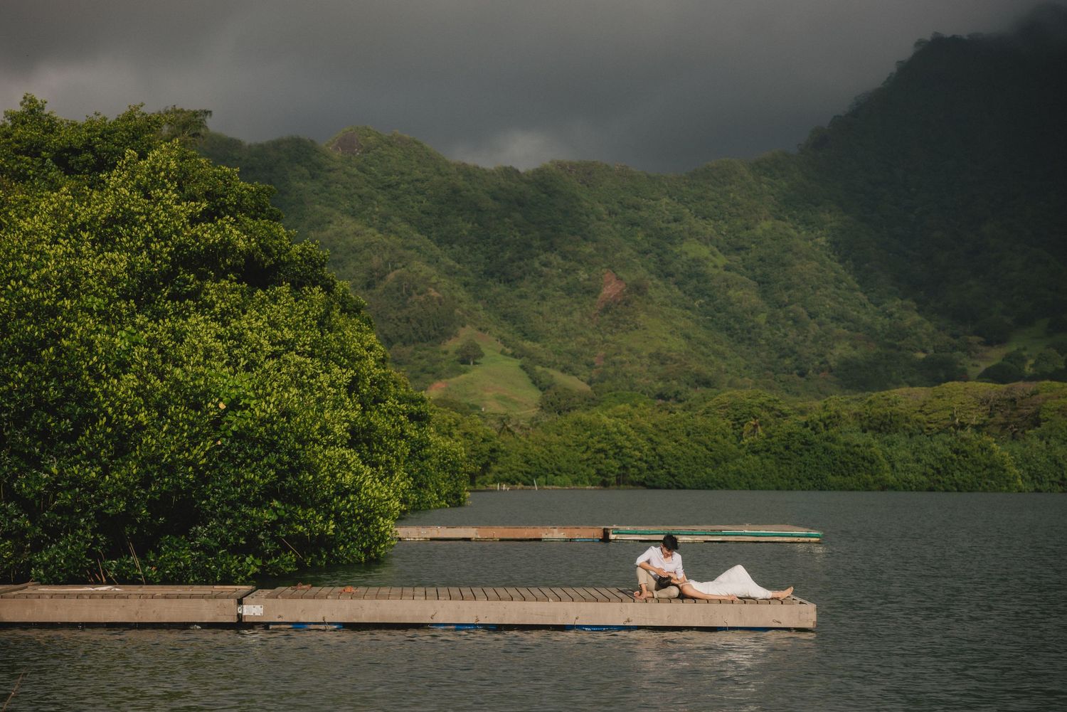 Couple sitting on a floating dock surrounded by lush green mountains and a calm lake under dramatic storm clouds.