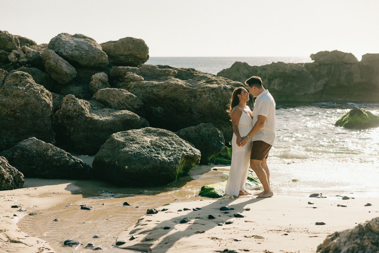 A couple embraces romantically on a sandy beach surrounded by large rocks with ocean waves in the background.
