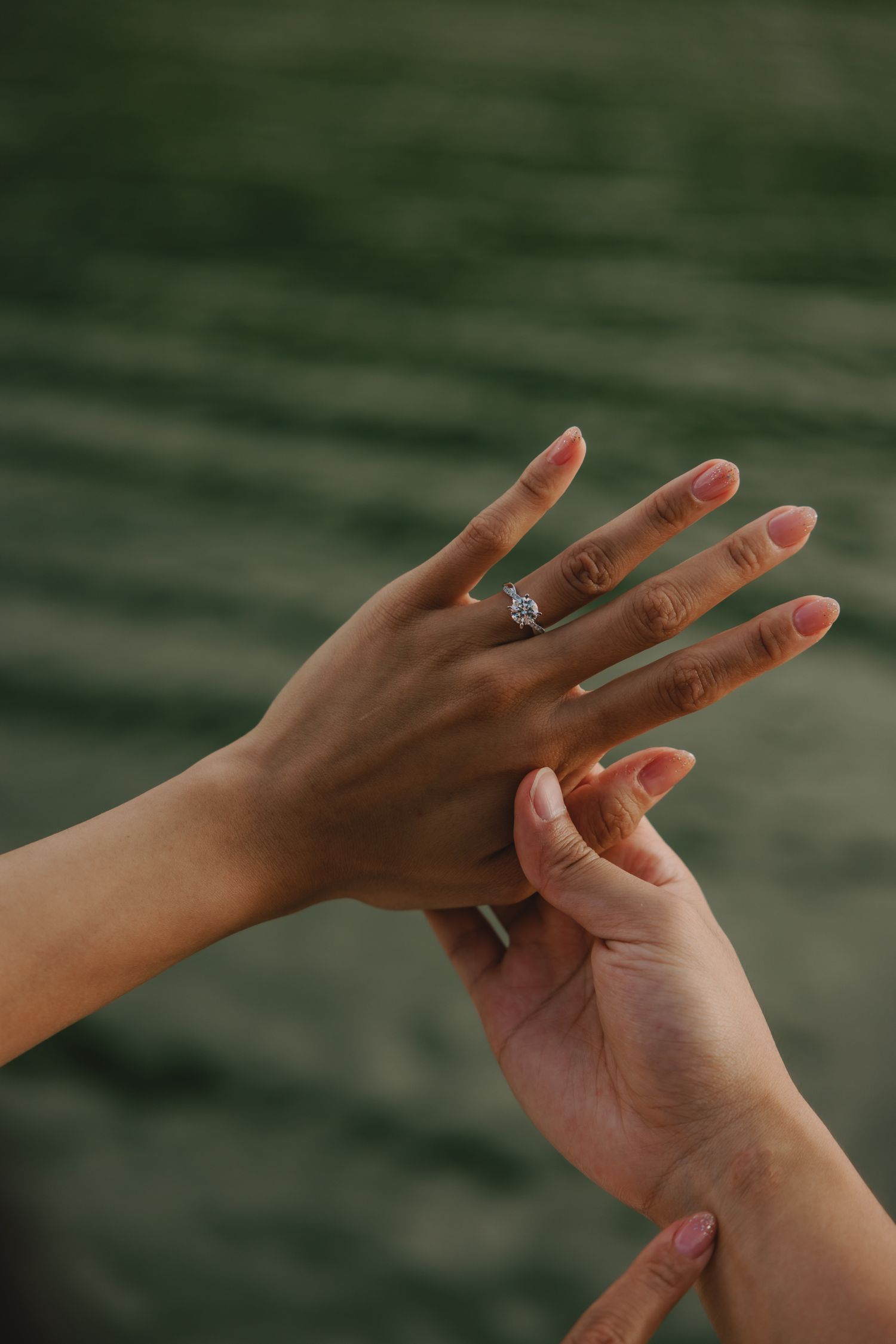 Two hands with an engagement ring being placed on a finger, with a blurred green water background.