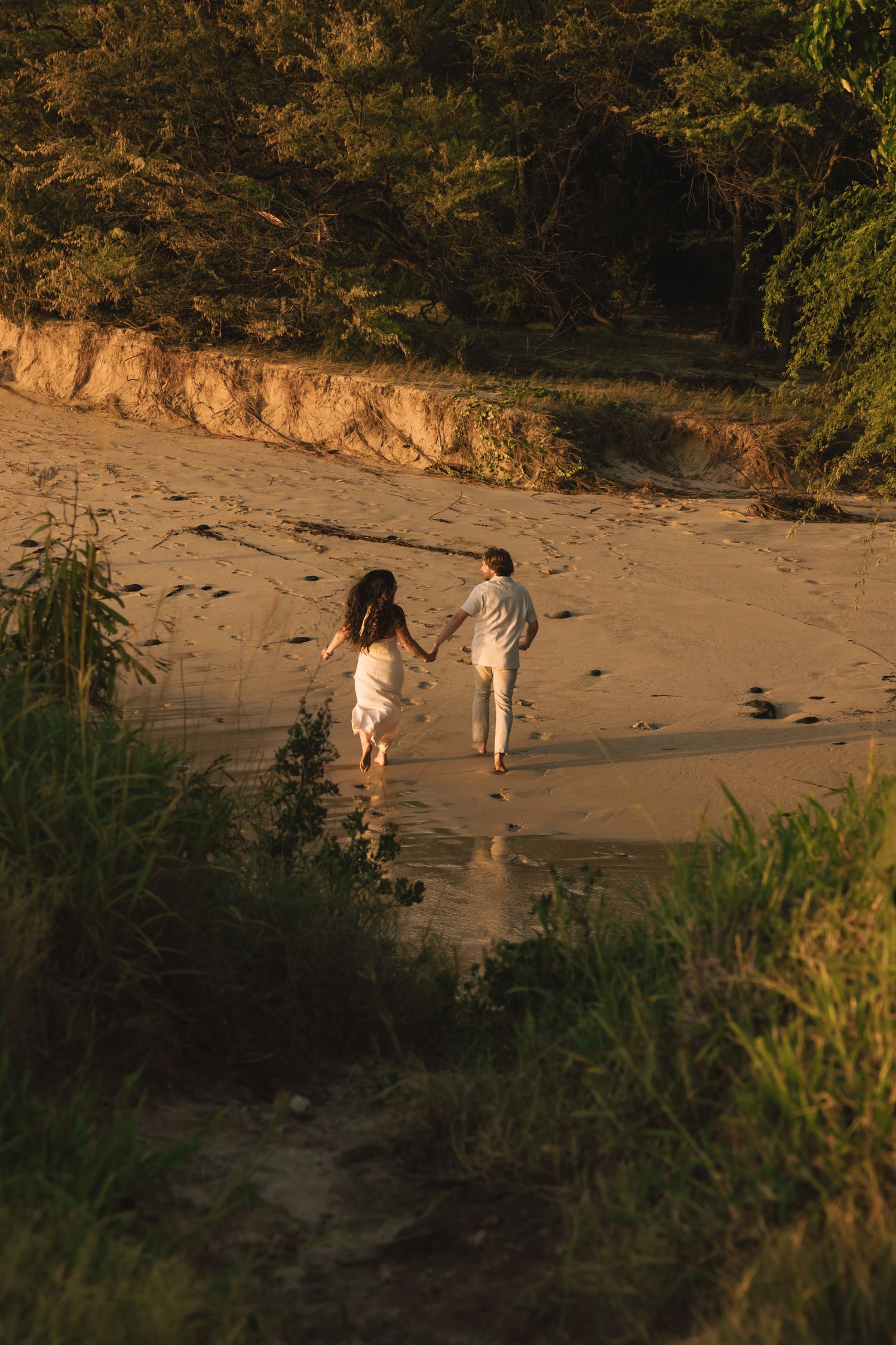 Couple holding hands walking on a sandy beach at golden hour with lush green trees in the background.