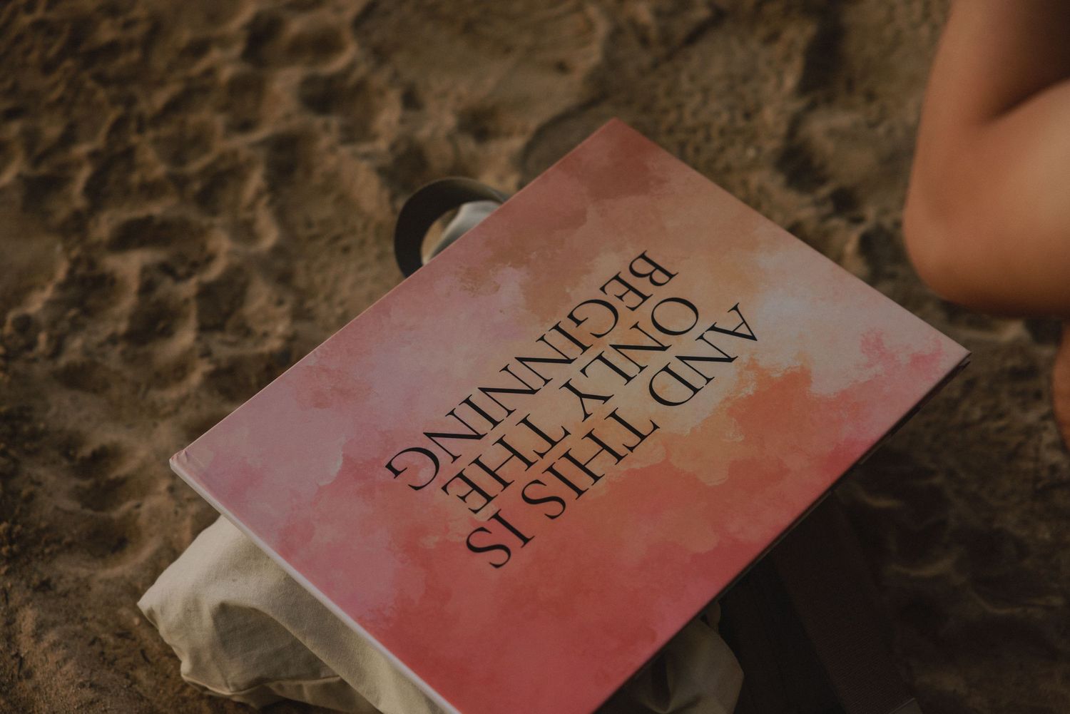 A pink watercolor book titled 'And This Is Only The Beginning' resting on sandy beach ground.