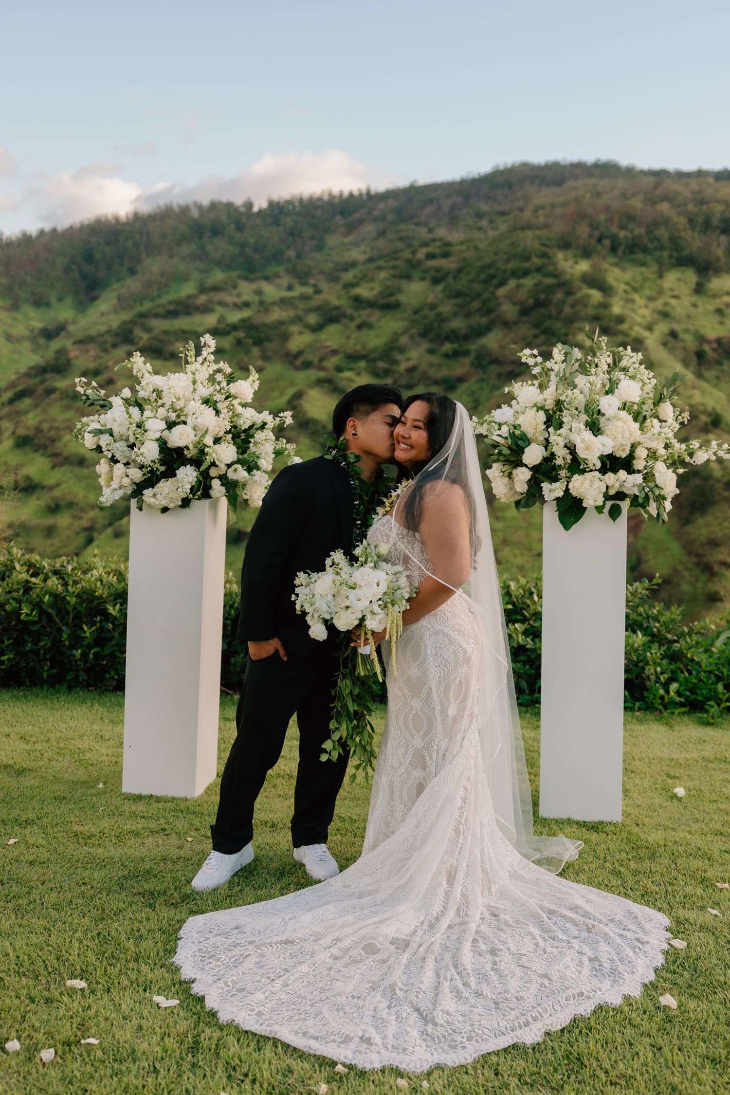 Bride and groom share a kiss at outdoor Hawaii wedding ceremony with white floral pillars and lush green mountains.