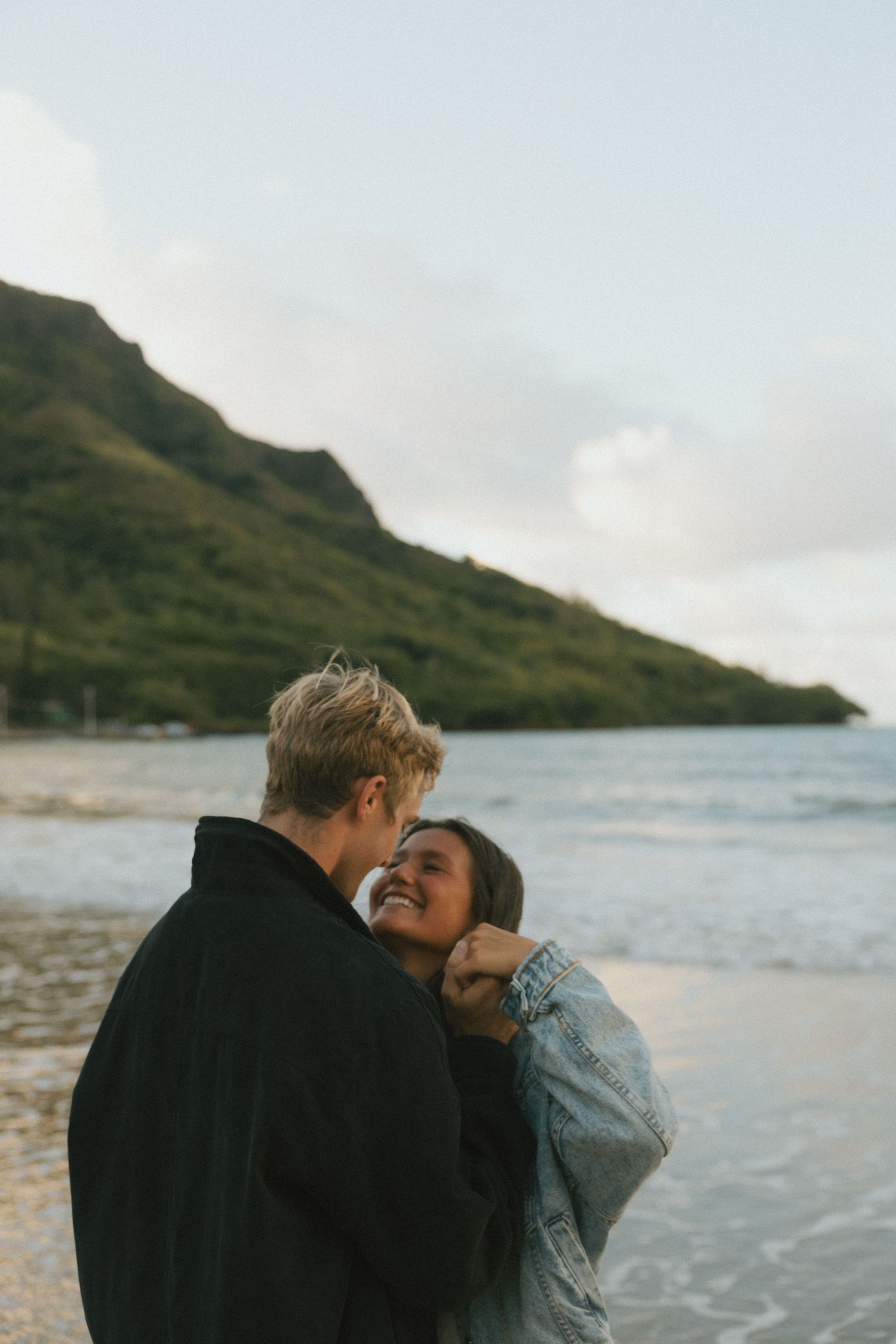 Couple embracing and laughing together on a beach with a mountain backdrop at sunset.