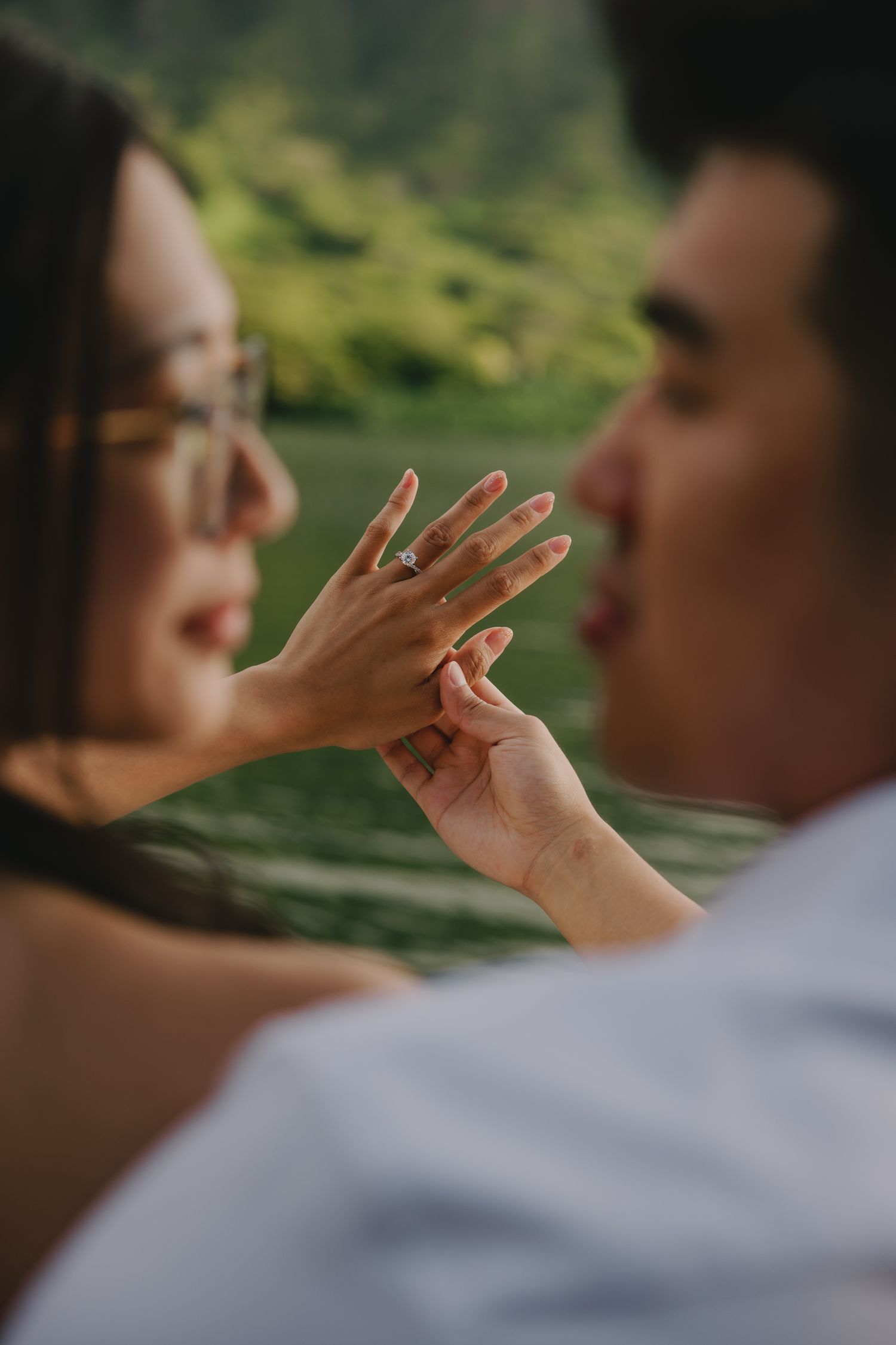 Couple sharing a romantic moment, woman displaying her engagement ring while smiling, with lush greenery in the background.